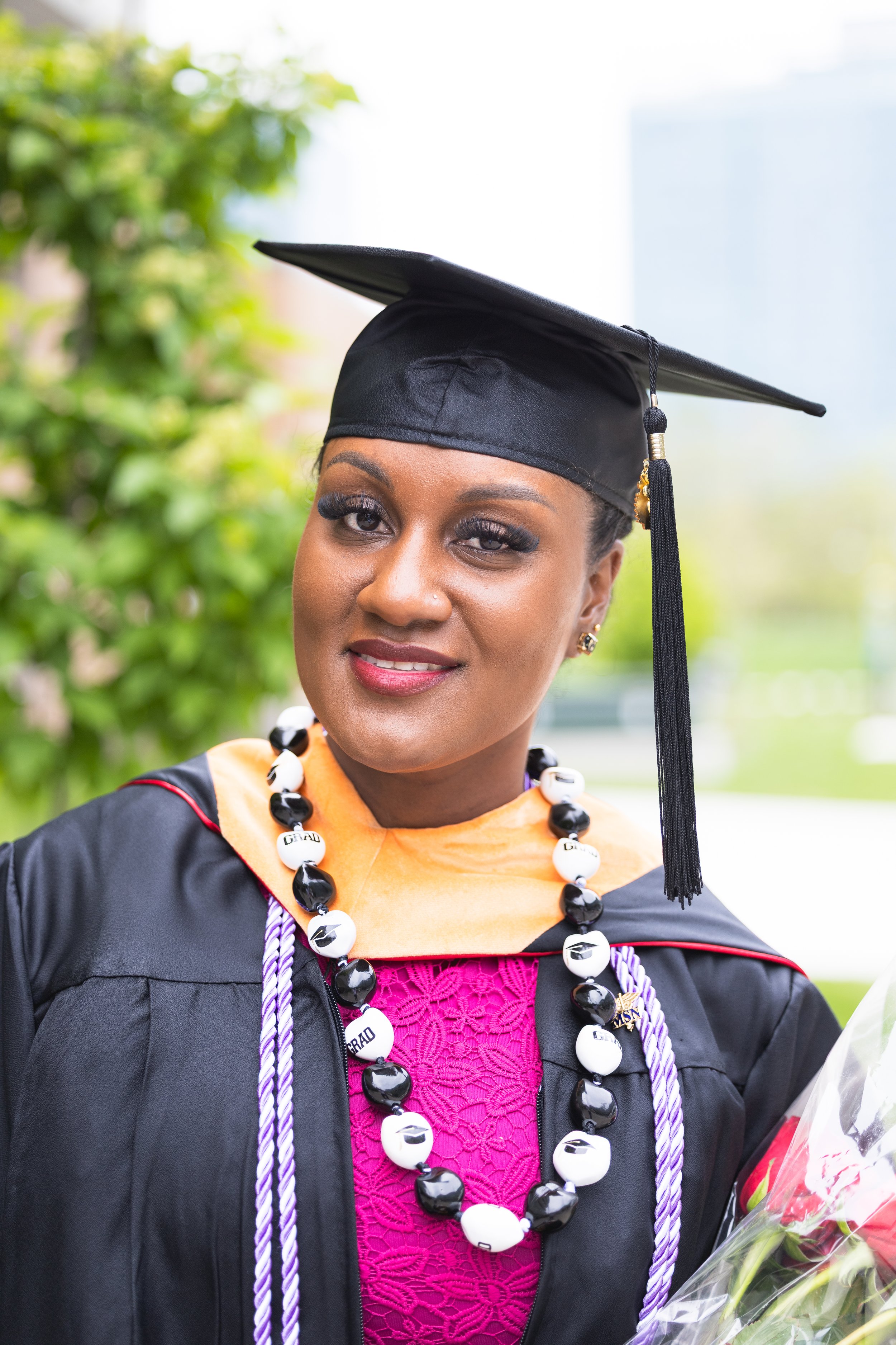 Young woman in graduation gown and cap, wearing graduation cords and beads, holding flowers, smiling outdoors.