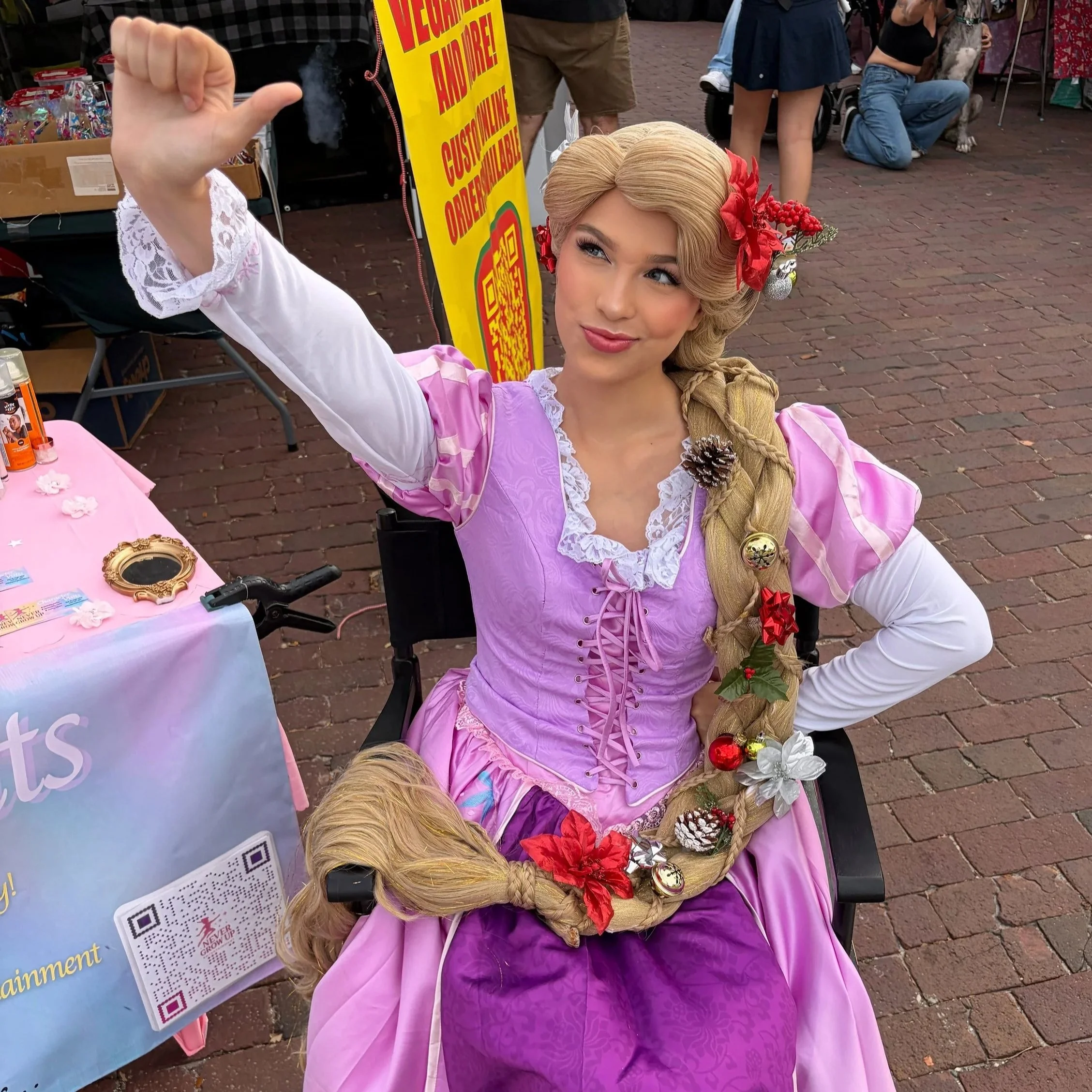 A woman dressed as Rapunzel with long braided hair decorated with Christmas ornaments and flowers, sitting at a table at an outdoor event, giving a thumbs-up gesture.