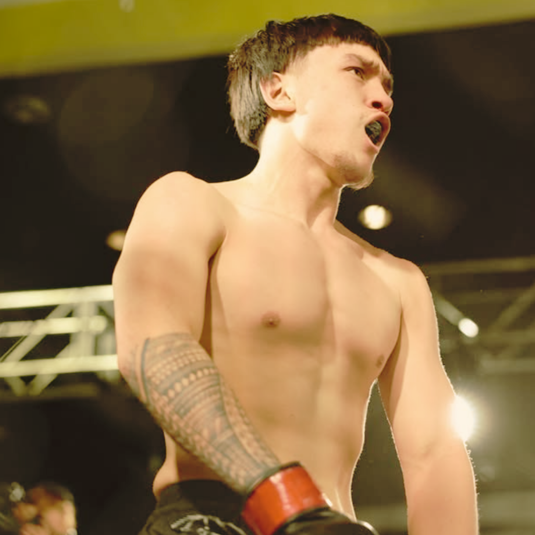 A shirtless male boxer with short dark hair and a tattooed arm, mid-action in a boxing ring with bright overhead lights.