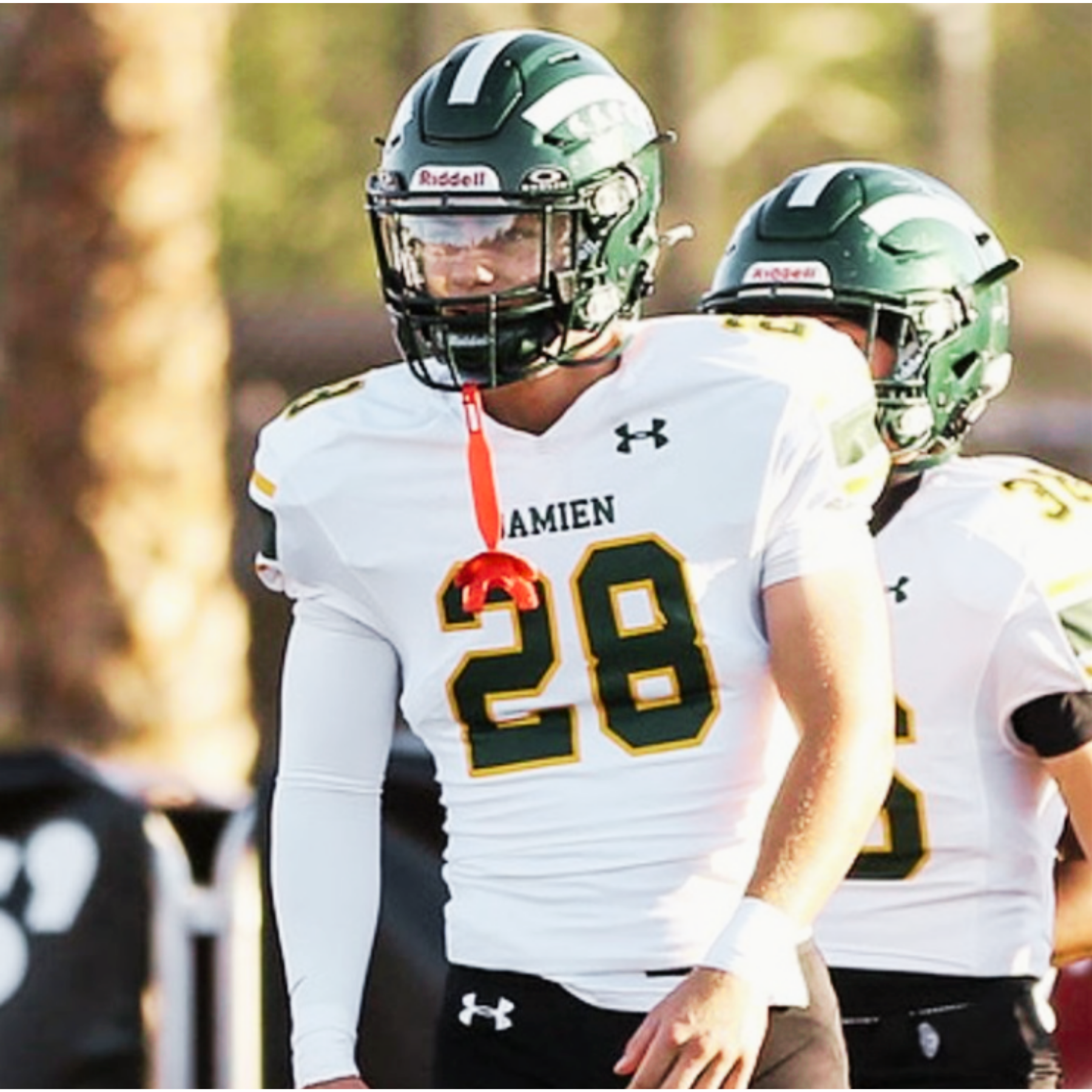 A football player wearing a green helmet and white jersey with black and yellow accents, standing on the field during a game. The player has the number 20 on the jersey and is part of a team with black shorts.