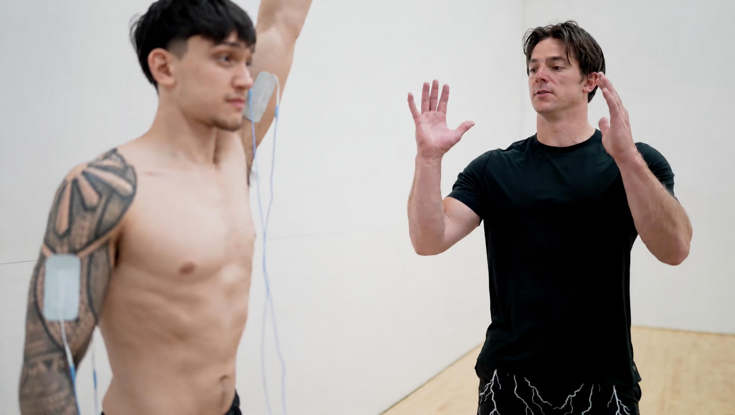 A shirtless man with tattoos on his arm and chest, wearing electrodes connected to his body, is being guided through an exercise by an instructor wearing a black t-shirt, in what appears to be a gym or therapy setting.