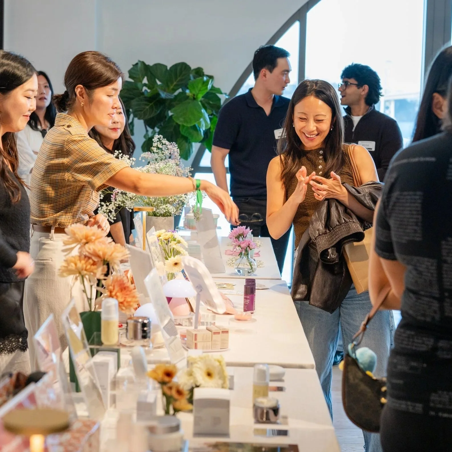 People shopping at a flower and beauty products display table during an indoor event, with floral arrangements and skincare items.