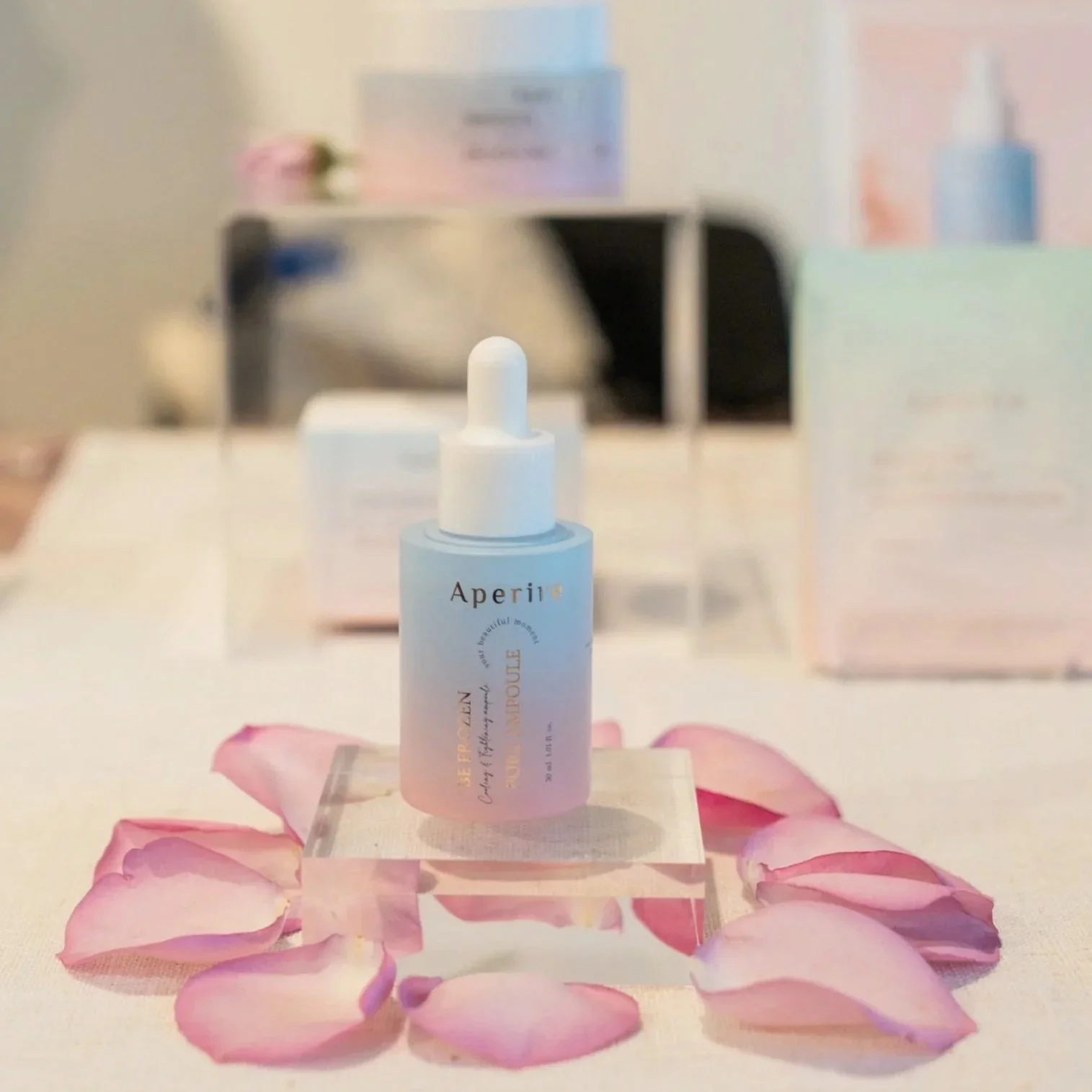 A skincare product bottle labeled 'Aperir' placed on a stand surrounded by pink rose petals, with a blurred background featuring a white desk, a pink box, and another skincare bottle.