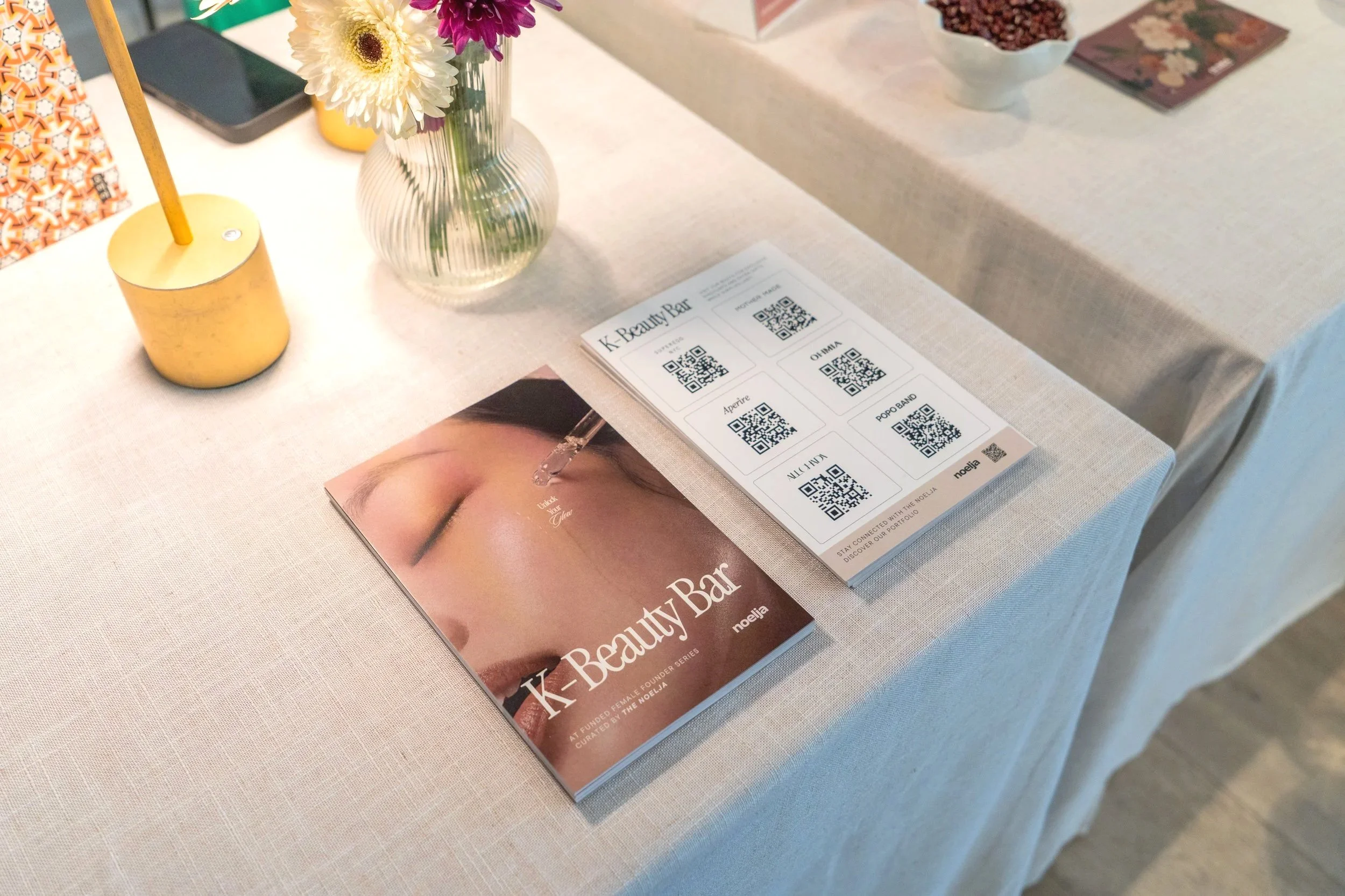 Table display at a beauty event featuring a brochure titled "K-Beauty Bar" with QR codes, a vase with white and purple flowers, a yellow candle holder, and a promotional magazine with a close-up of a woman's eye and eyebrow.