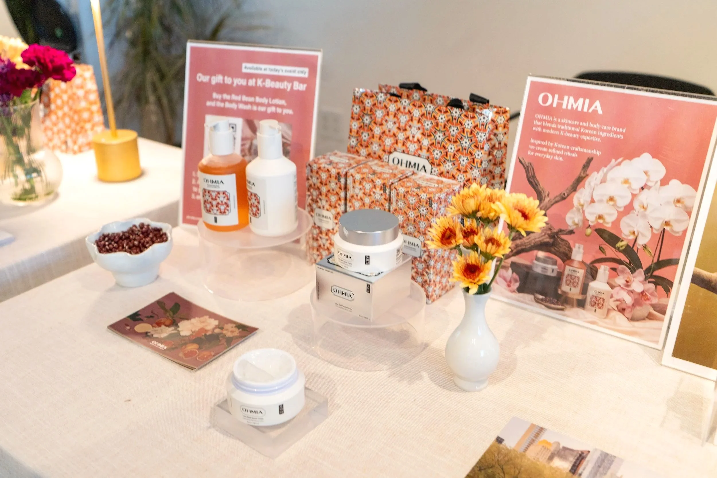 Display table featuring OHMIA skin and body care products, promotional signs, flowers, and product packaging at a beauty event.