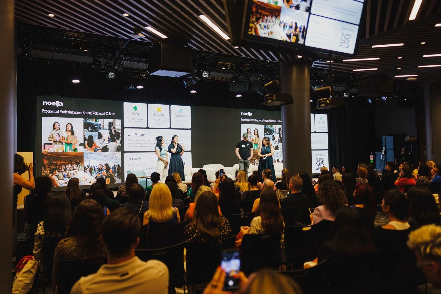 A conference or seminar with a large audience facing a stage with four speakers. The stage features a large digital screen displaying presentation slides about a company called noelja, focusing on marketing across beauty, wellness, and culture.