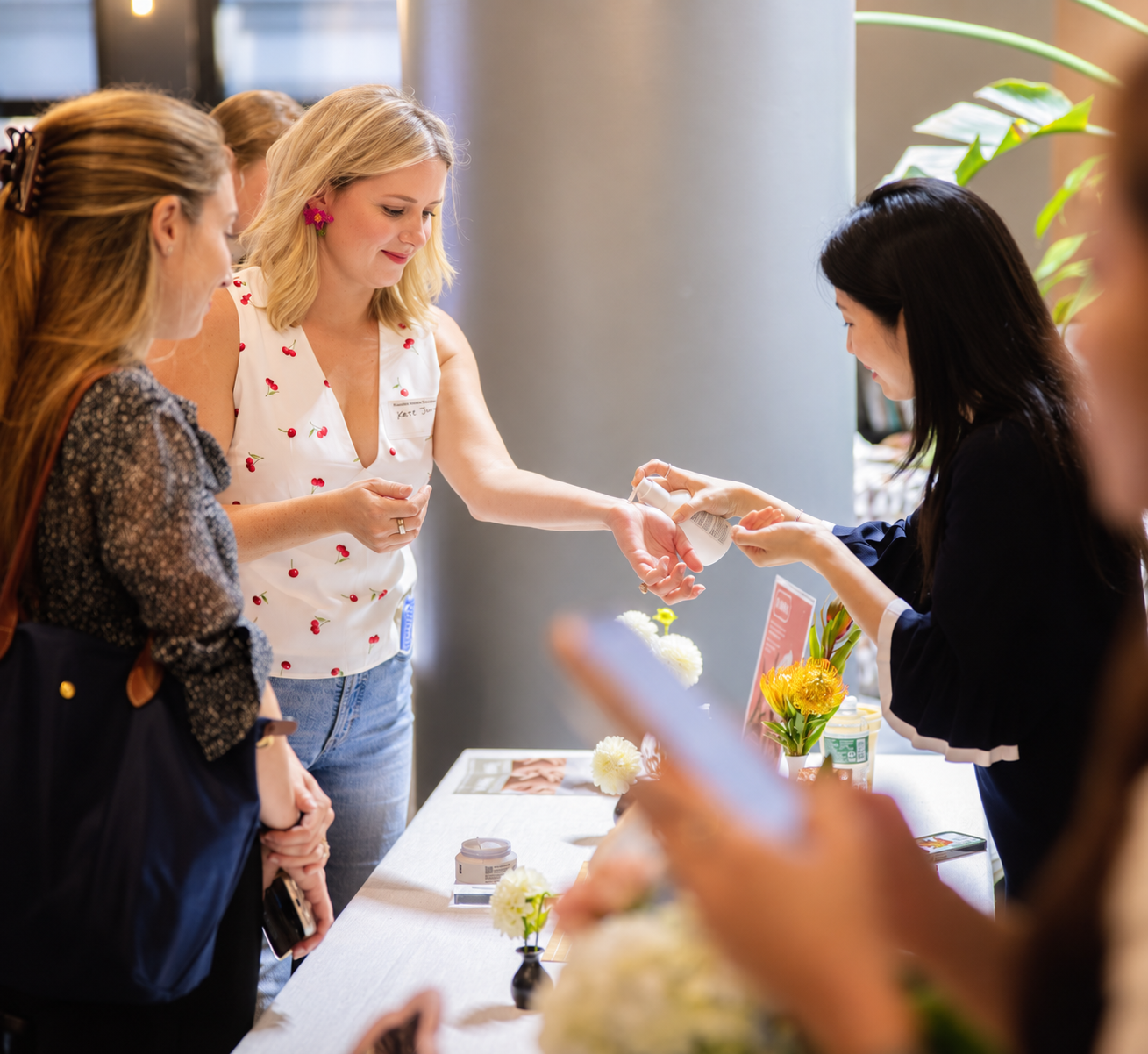 Woman at a table handing out skincare samples to a line of women in a bright, modern space decorated with flowers.