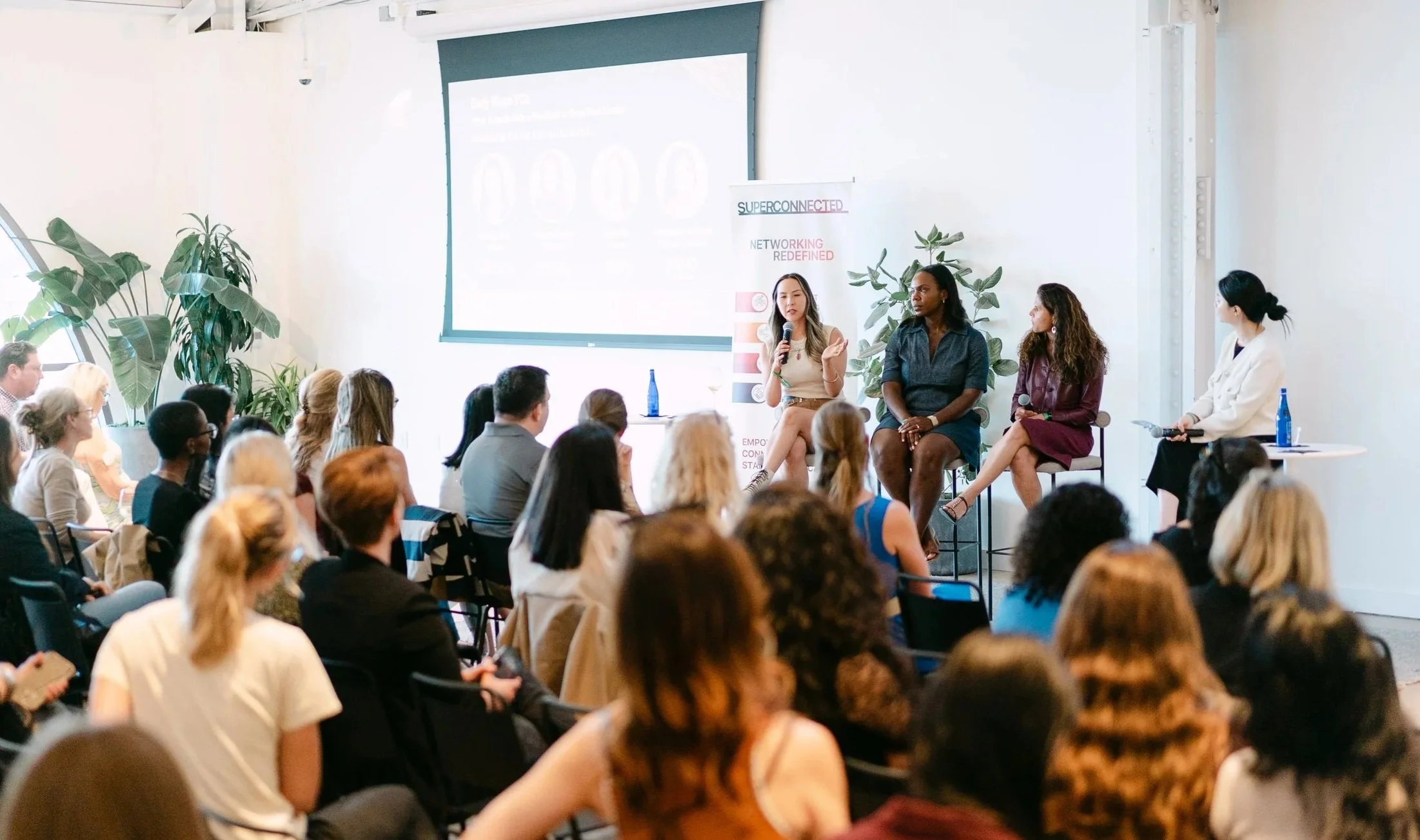 A panel discussion at a conference with four women speakers seated in front of an audience. There are large plants on either side of the speakers and a presentation screen behind them displaying a slide. The audience is attentively listening.