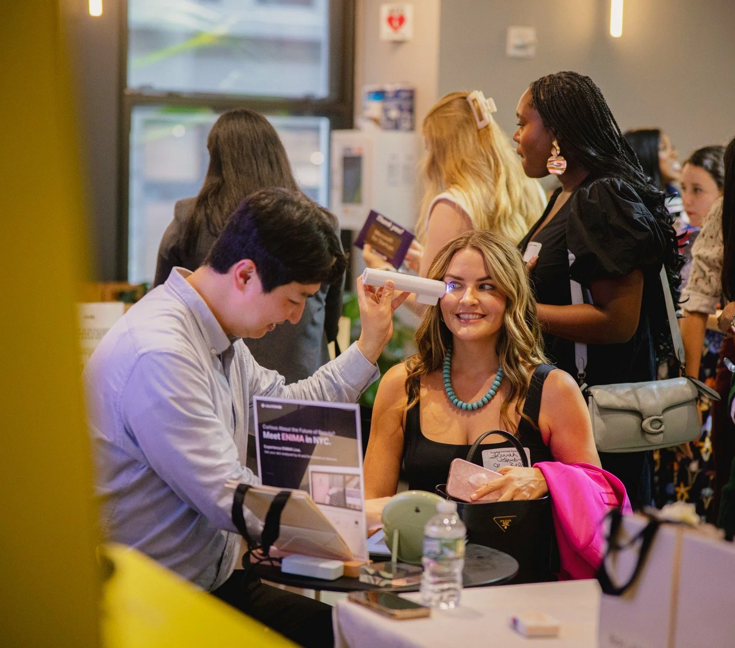 A woman sitting at a table getting her eyes checked by a young male optometrist at an indoor event.