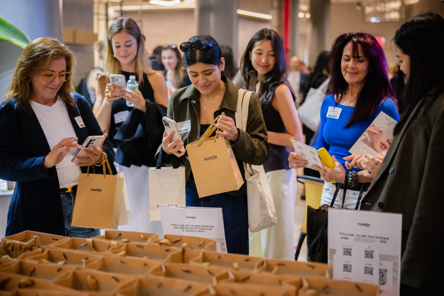 Group of women shopping at an indoor event, examining products and using their phones, standing around a display table with small bags and signs.