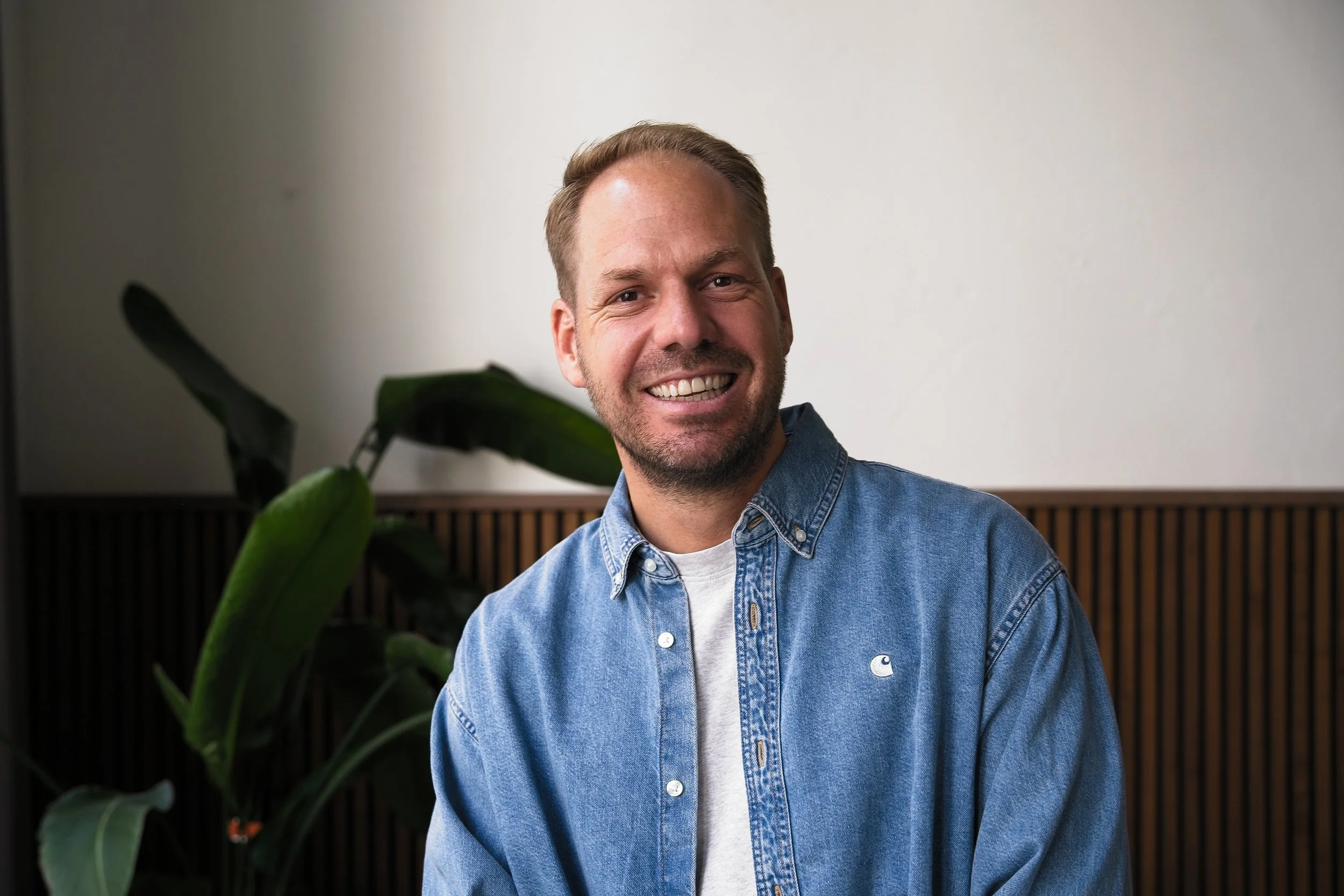 Een lachende man met kort haar en een lichte baard, gekleed in een blauwe spijkerjas en een wit T-shirt, zittend voor een neutrale achtergrond met een groene plant op de voorgrond.