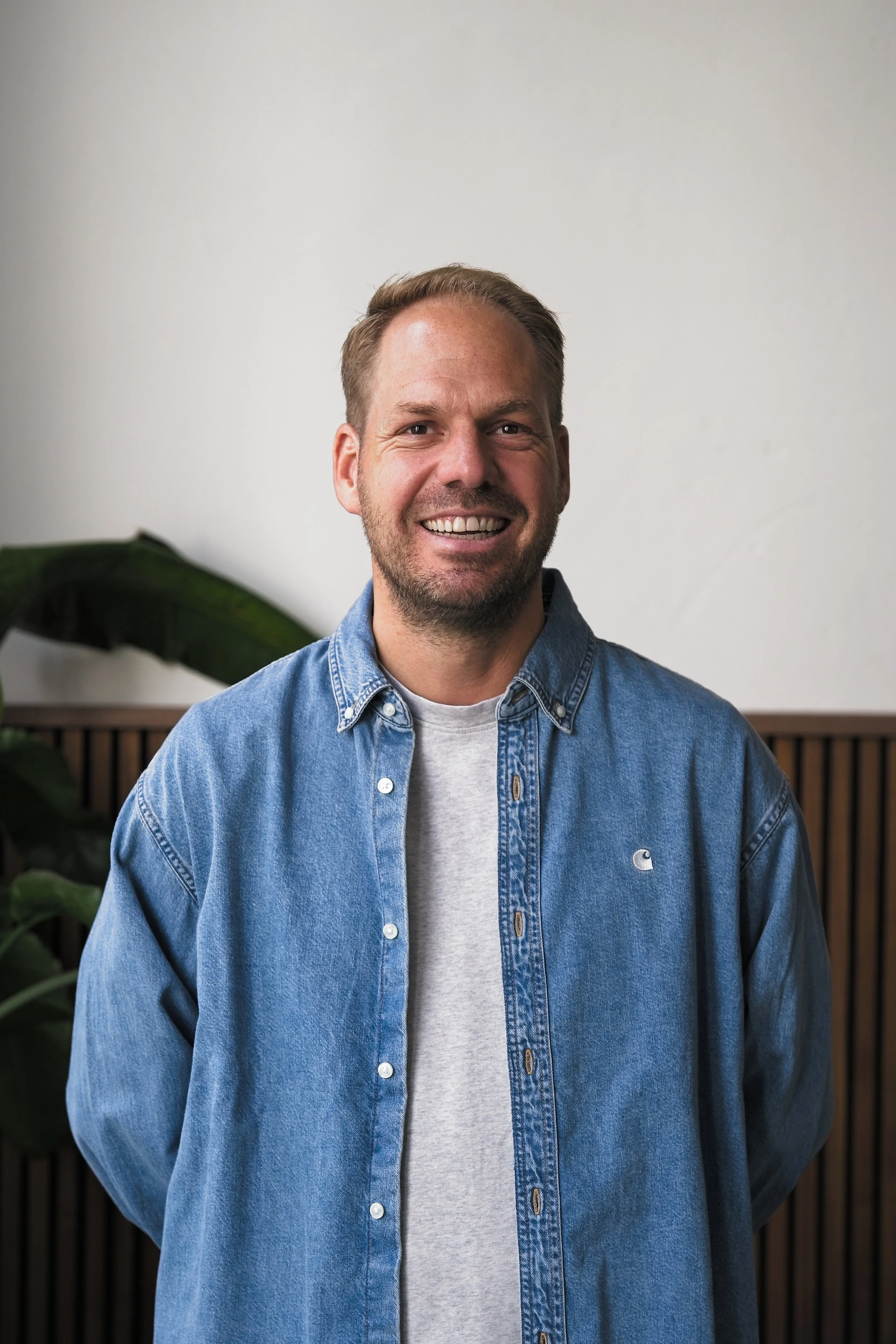 Lachende man met korte haardracht draagt een lichtblauwe denim overhemd en lichtgrijze T-shirt, staat binnen voor een witte muur en houten paneel, met groene plant op de achtergrond.
