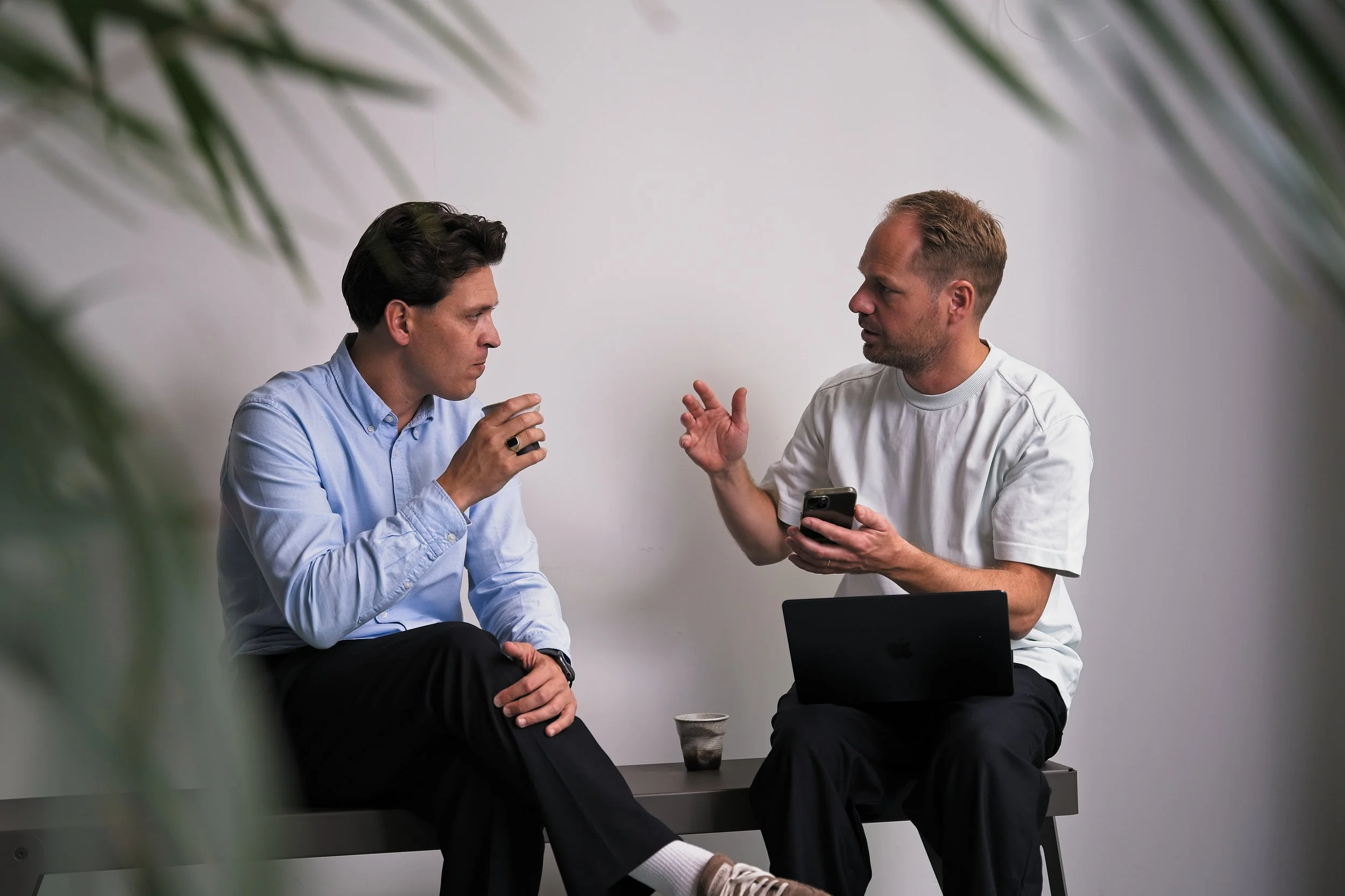 Twee mannen zitten op een bankje in gesprek, de man aan de rechterkant toont op zijn mobiele telefoon en heeft een laptop op zijn schoot, terwijl de man aan de linkerkant aandachtig luistert.