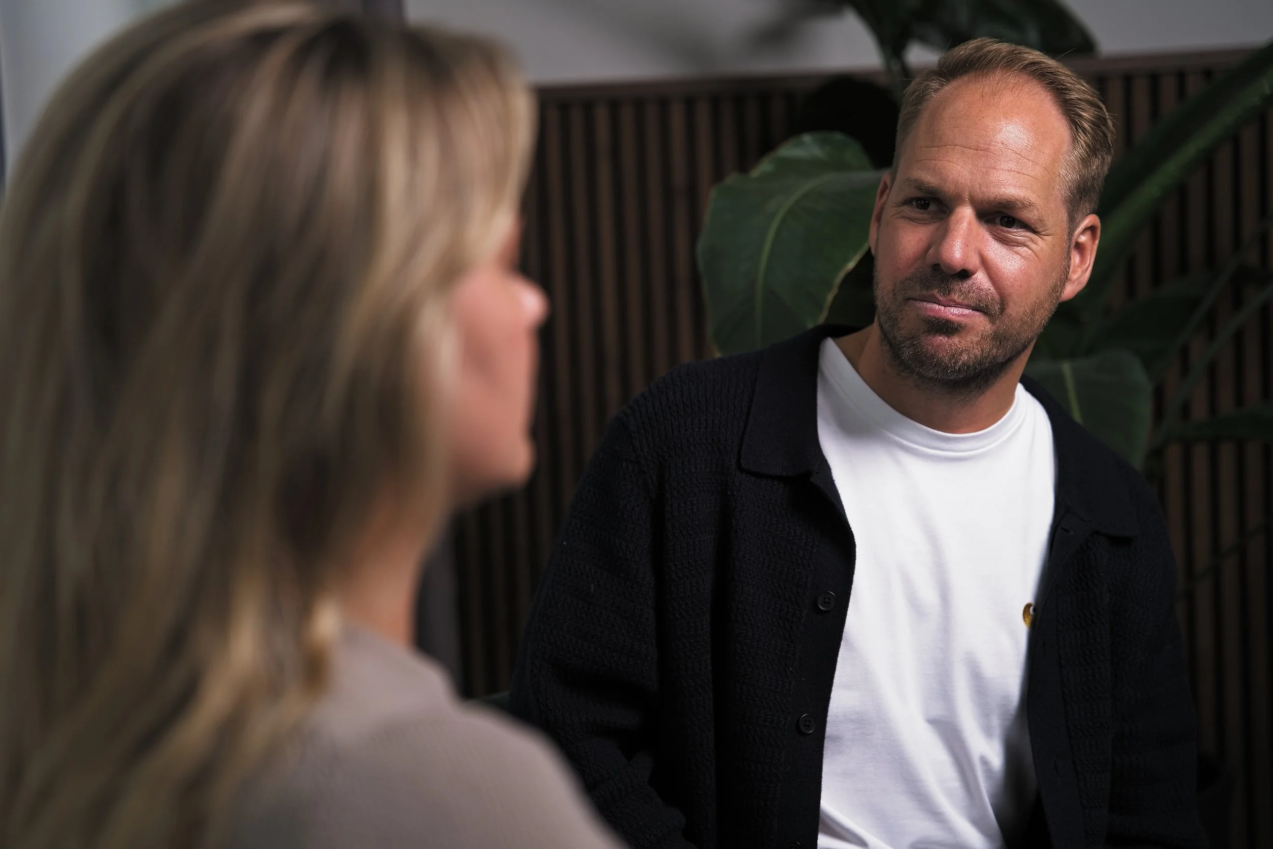 Een man en een vrouw hebben een gesprek in een interieur met grote groene planten op de achtergrond.