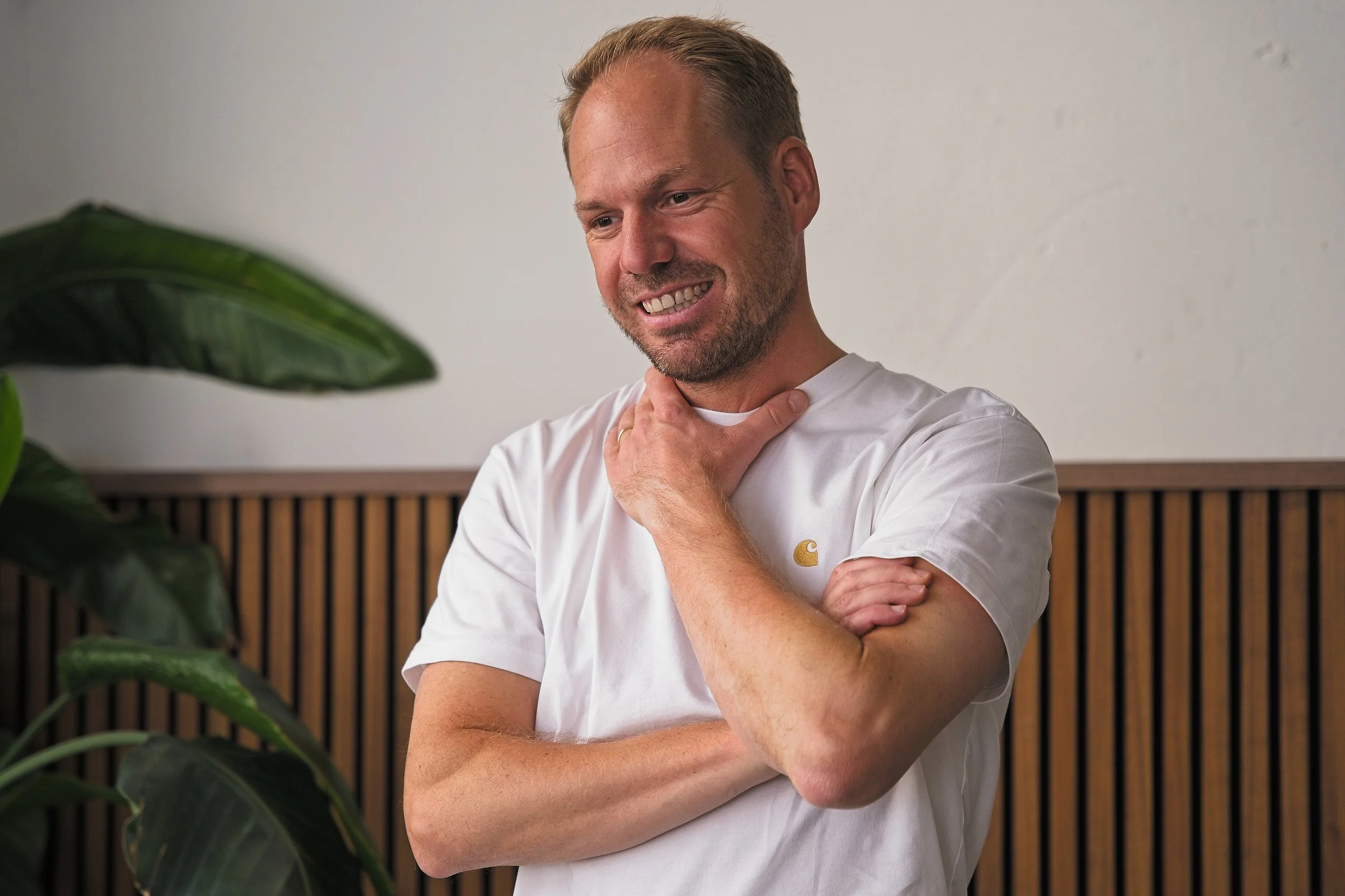 Man met korte haar, lichte huid, witte T-shirt, glimlach, staat binnen in een kamer met houten lamellen achterwand, naast een groene plant