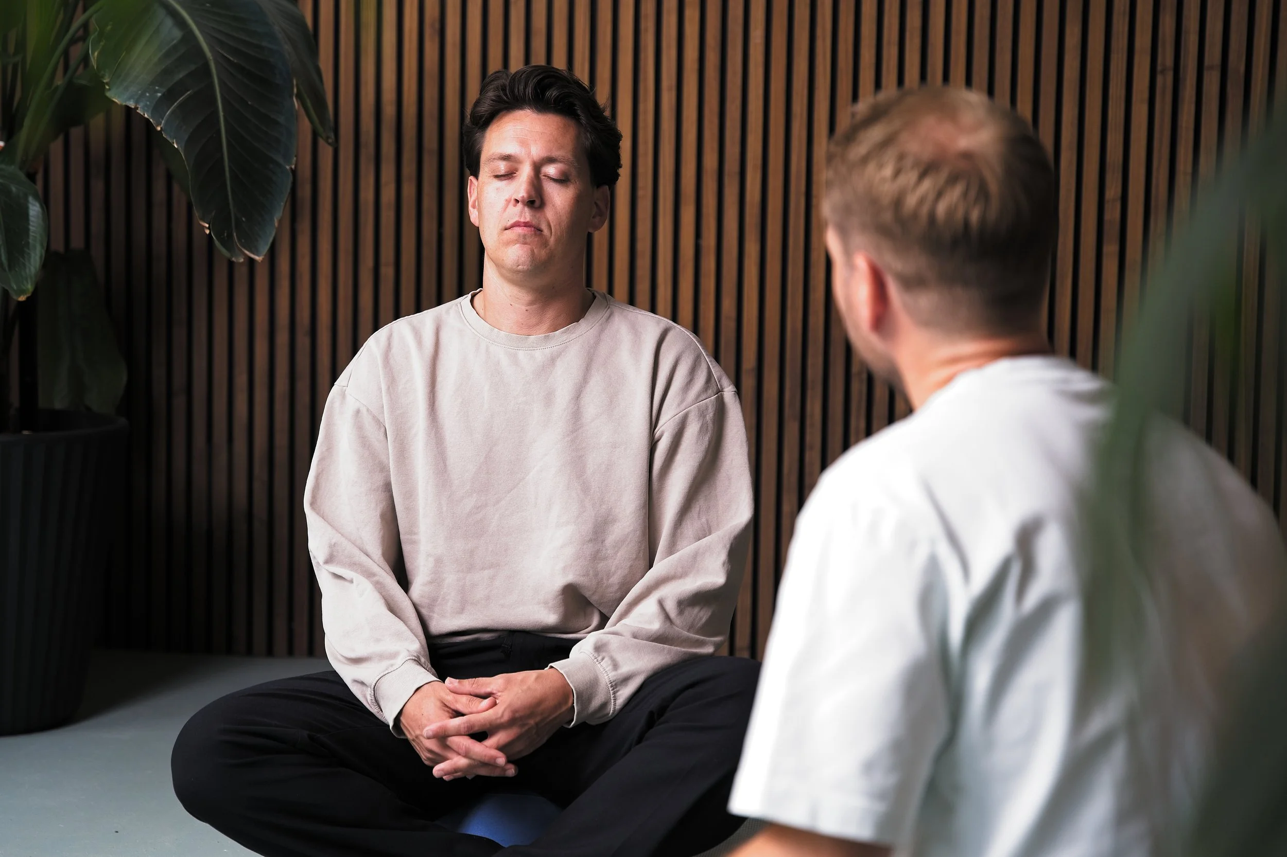 Twee mannen zitten op de grond, de een met gesloten ogen, in een kamertje met houten wand en planten.