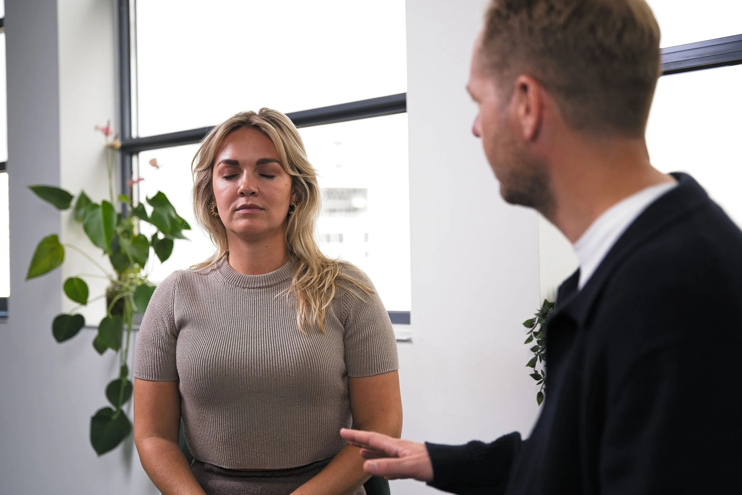 Een man en een vrouw in een kantoor, de vrouw heeft haar ogen dicht en lijkt te mediteren of te concentreren, terwijl de man met zijn hand gebaren maakt tijdens een gesprek.