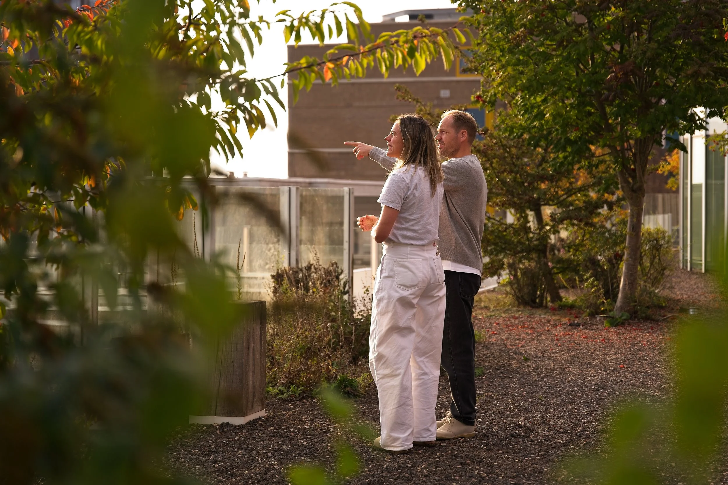 Twee mensen staan in een tuin of park, een vrouw die naar de rechterkant wijst en een man die naar haar kijkt, omringd door bomen en struiken, met een gebouw op de achtergrond.
