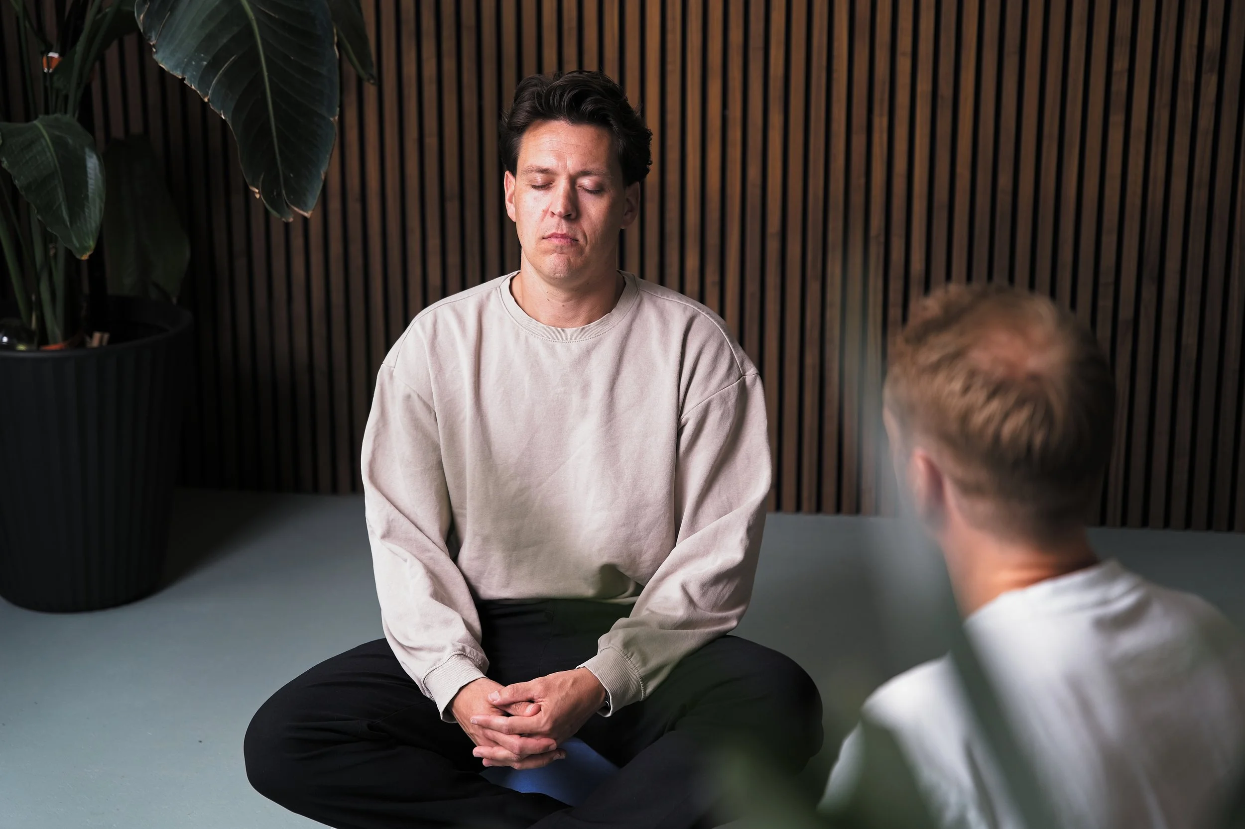 Twee mannen zitten op de vloer en hebben een gesprek. De man aan de voorkant heeft zijn ogen dicht en lijkt rustig. De man op de achtergrond heeft zijn rug naar de camera. De achtergrond bestaat uit een houten wand en er staat een grote plant links in de hoek.