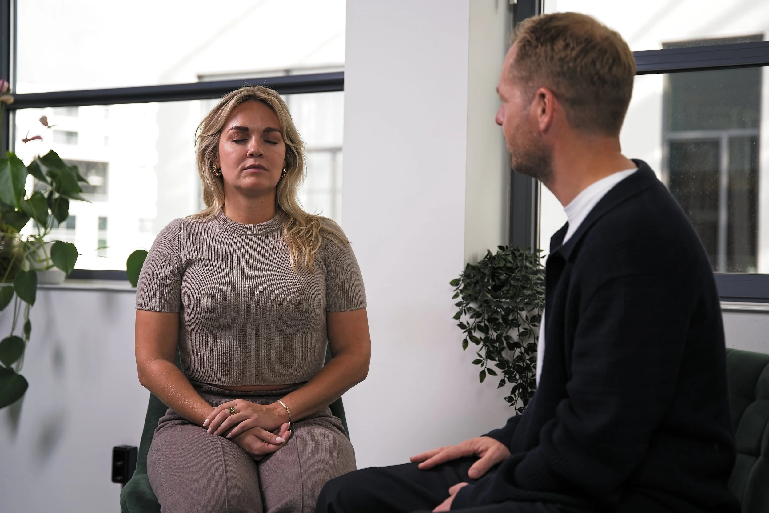 Een vrouw zit met gesloten ogen in een gespreksruimte met een man die tegenover haar zit. Ze lijken in meditatie of diepe gedachten. Er zijn planten en ramen op de achtergrond.