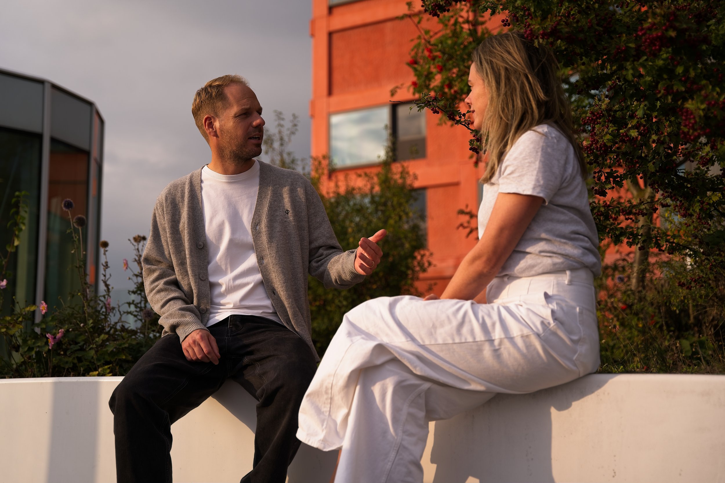 Man en vrouw zitten op een muur en praten buiten met een groene achtergrond en een rood gebouw op de achtergrond, onder een bewolkte hemel.