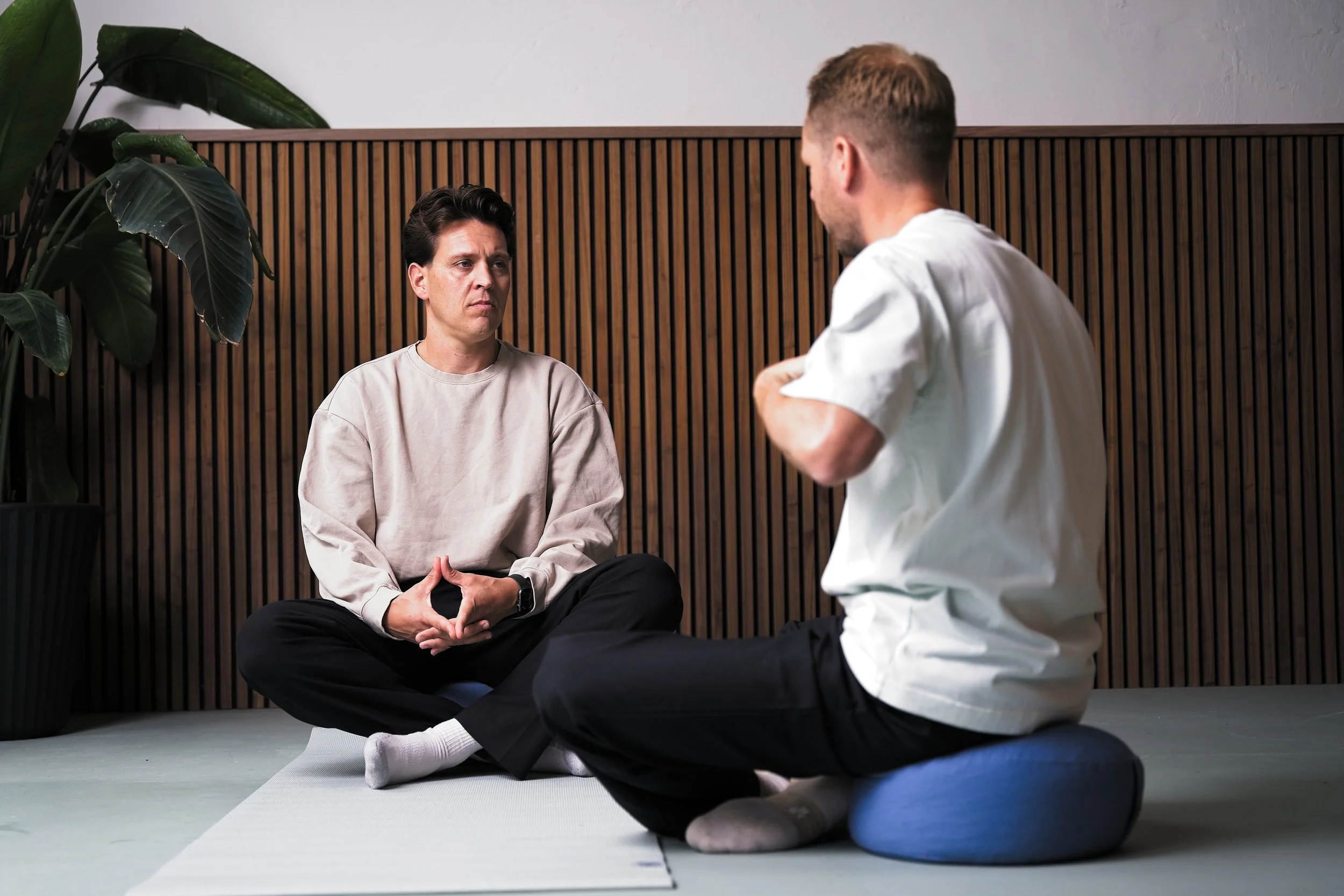 Twee mensen zitten op de grond in een gespreksruimte, een vrouw kijkt serieus terwijl een man met een witte T-shirt op een meditatiekussen zit.