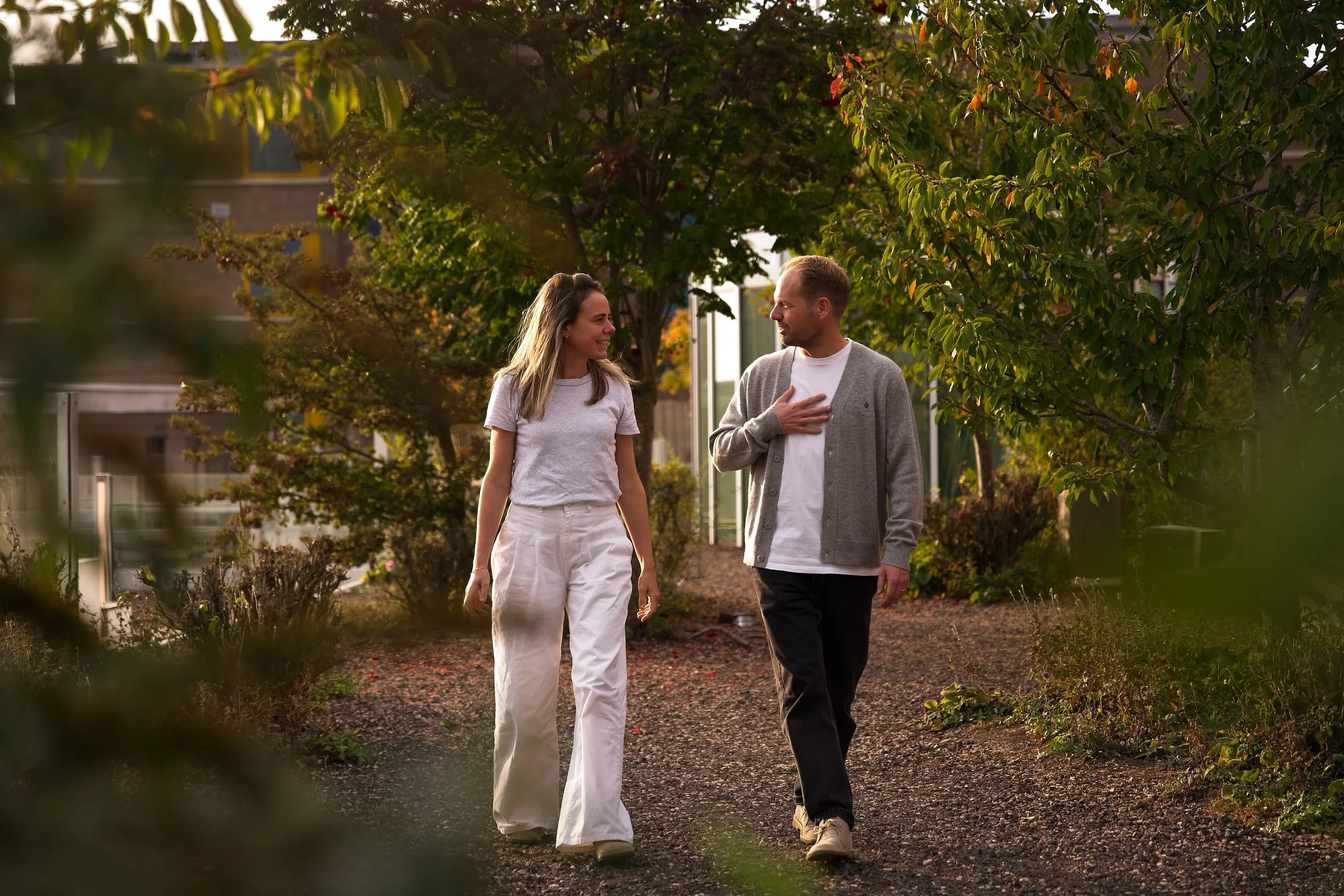 Een man en vrouw wandelen samen door een tuin, ze praten en lachen. De vrouw draagt een wit T-shirt en witte broek, de man draagt een grijze cardigan en zwarte broek.
