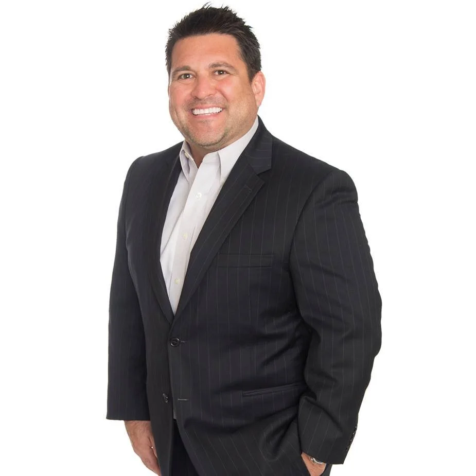 A smiling man wearing a dark pinstripe suit and a white dress shirt, standing against a plain white background.