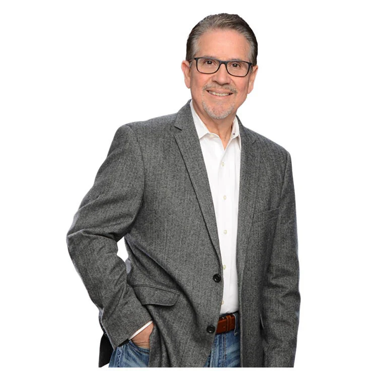 A middle-aged man with gray hair and glasses smiling, wearing a gray blazer over a white shirt, standing with one hand in his pocket against a white background.