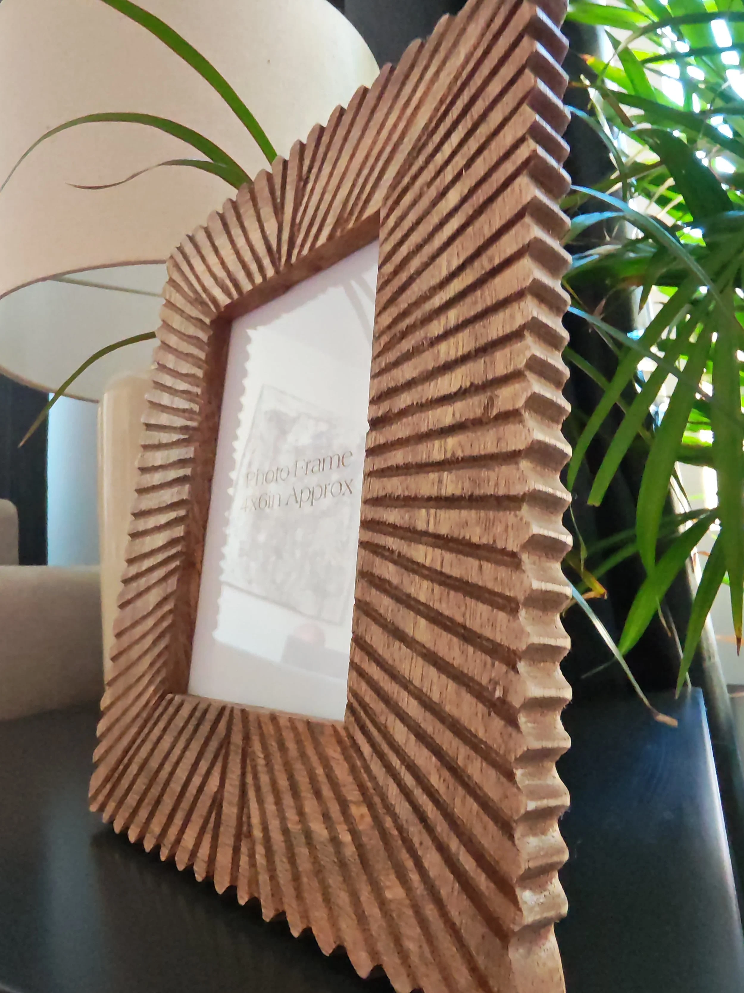 Decorative wooden photo frame with steps design, placed on a black surface, surrounded by green houseplants and a beige lamp in the background.
