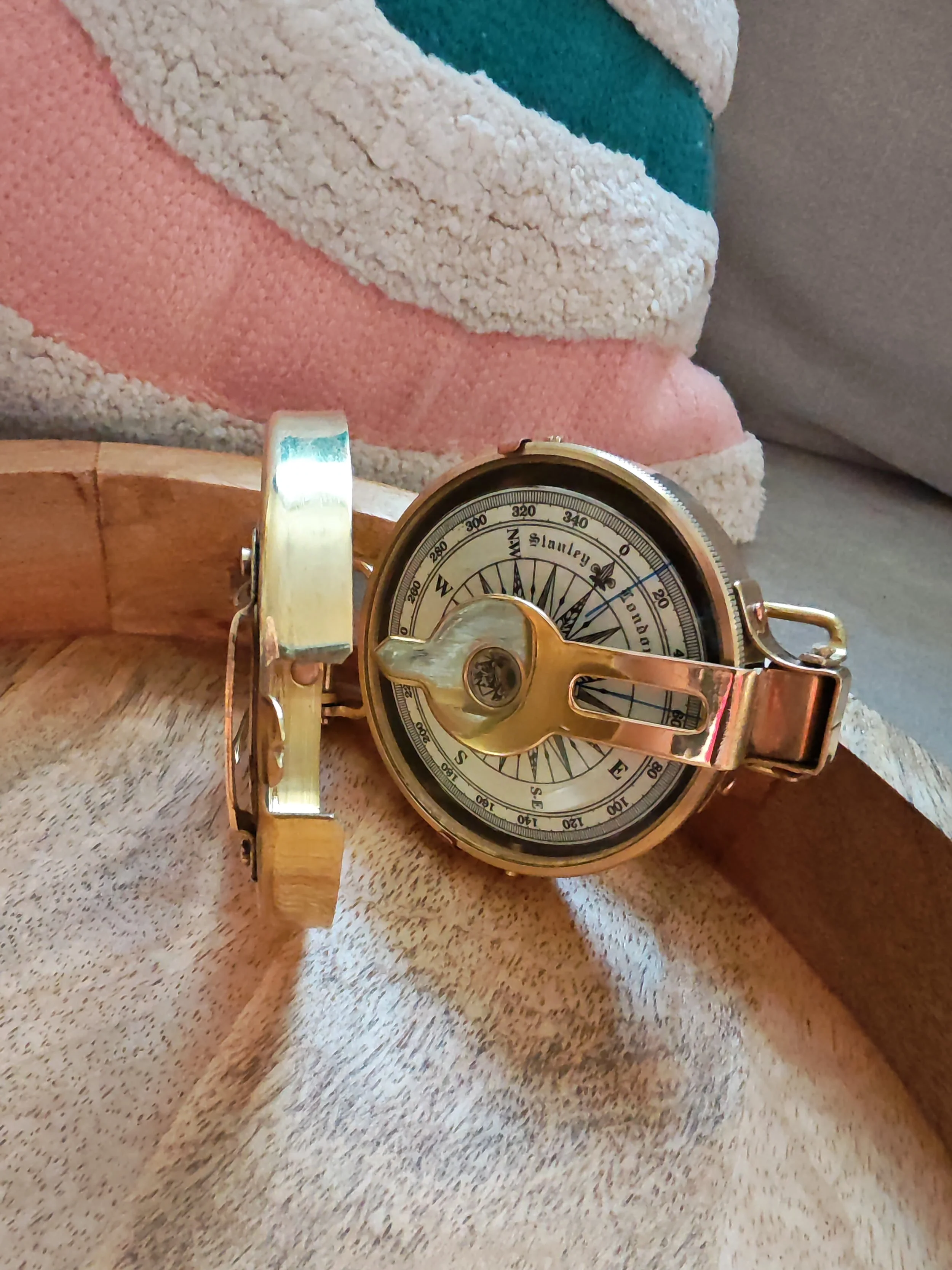 A vintage compass with a gold-colored metal case and band resting on a light-colored fabric surface, with a pink, blue, and cream striped blanket in the background.