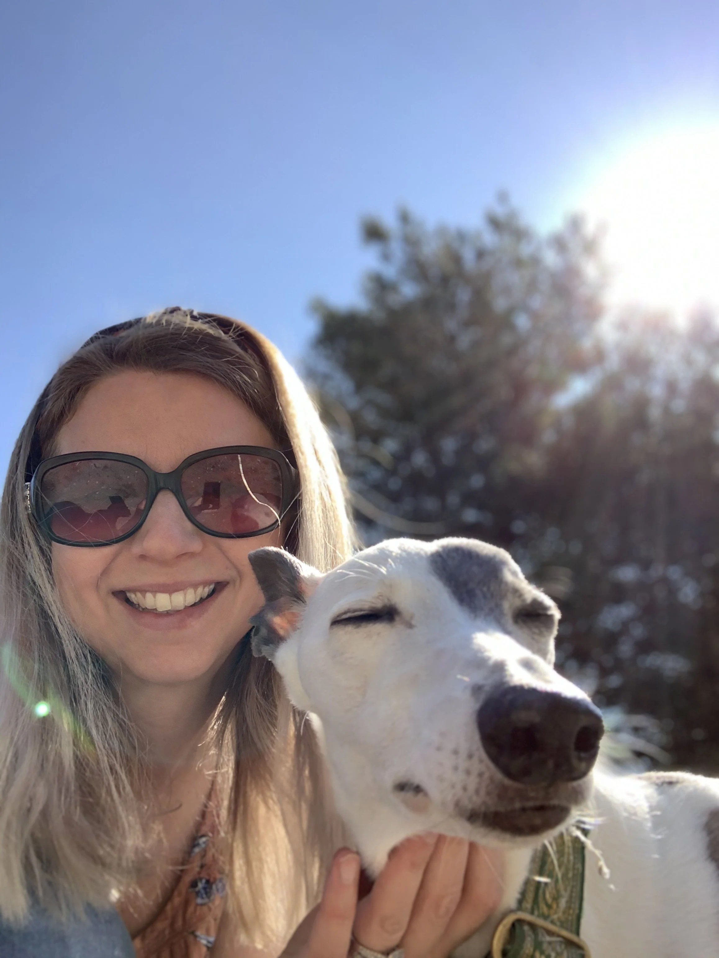 A woman with long blonde hair, wearing large sunglasses and a smile, takes a selfie with a smiling white and gray dog outdoor on a sunny day with a blue sky and trees in the background.