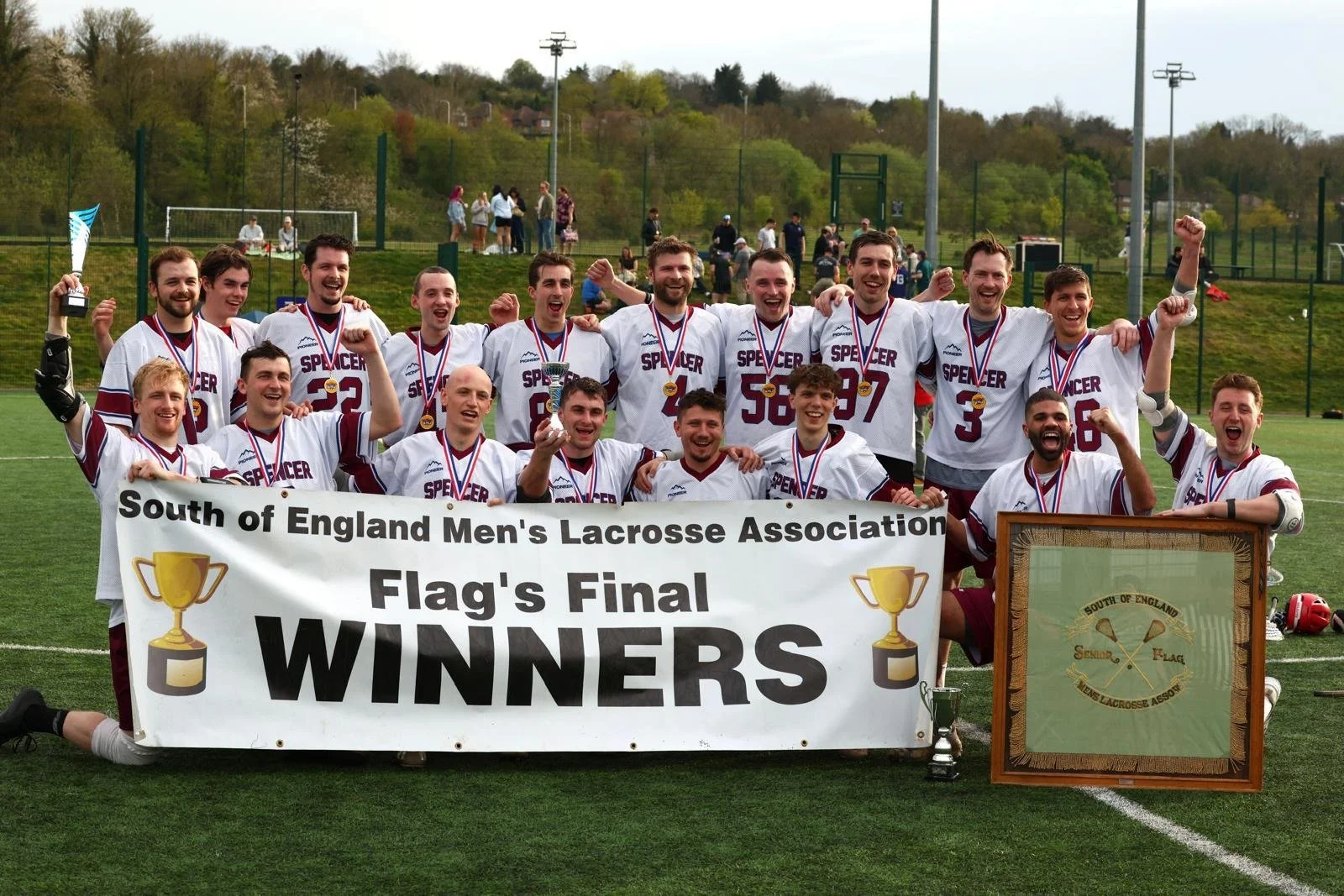 A group of lacrosse players in white jerseys celebrating after winning a championship. They are holding a banner that reads 'South of England Men's Lacrosse Association Flag's Final WINNERS' with two gold trophies. The players are smiling, cheering, and some are raising fists. They are on a sports field with a green background and spectators in the distance.