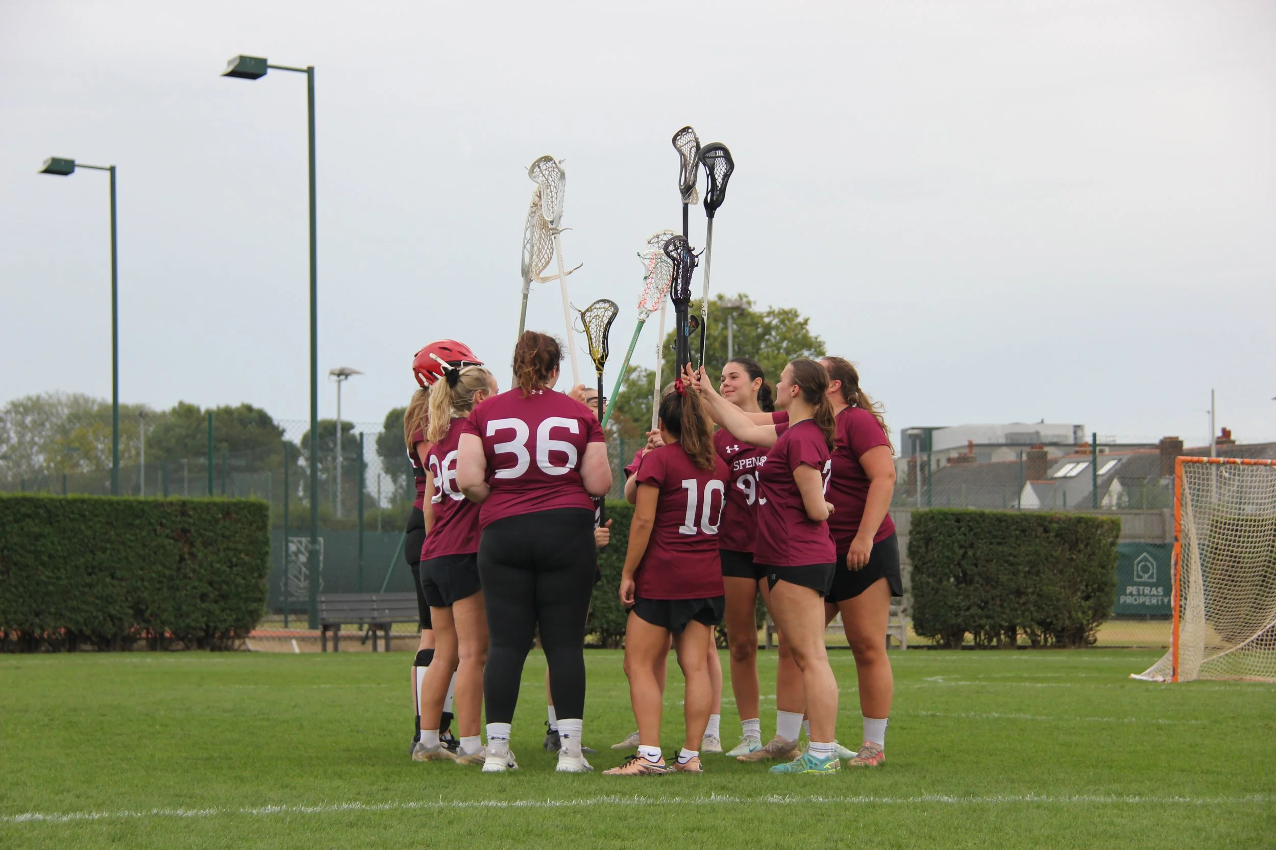 A group of female lacrosse players wearing maroon jerseys and black shorts gathered together on a field, holding lacrosse sticks with the players in the center apparently celebrating or having a team huddle.