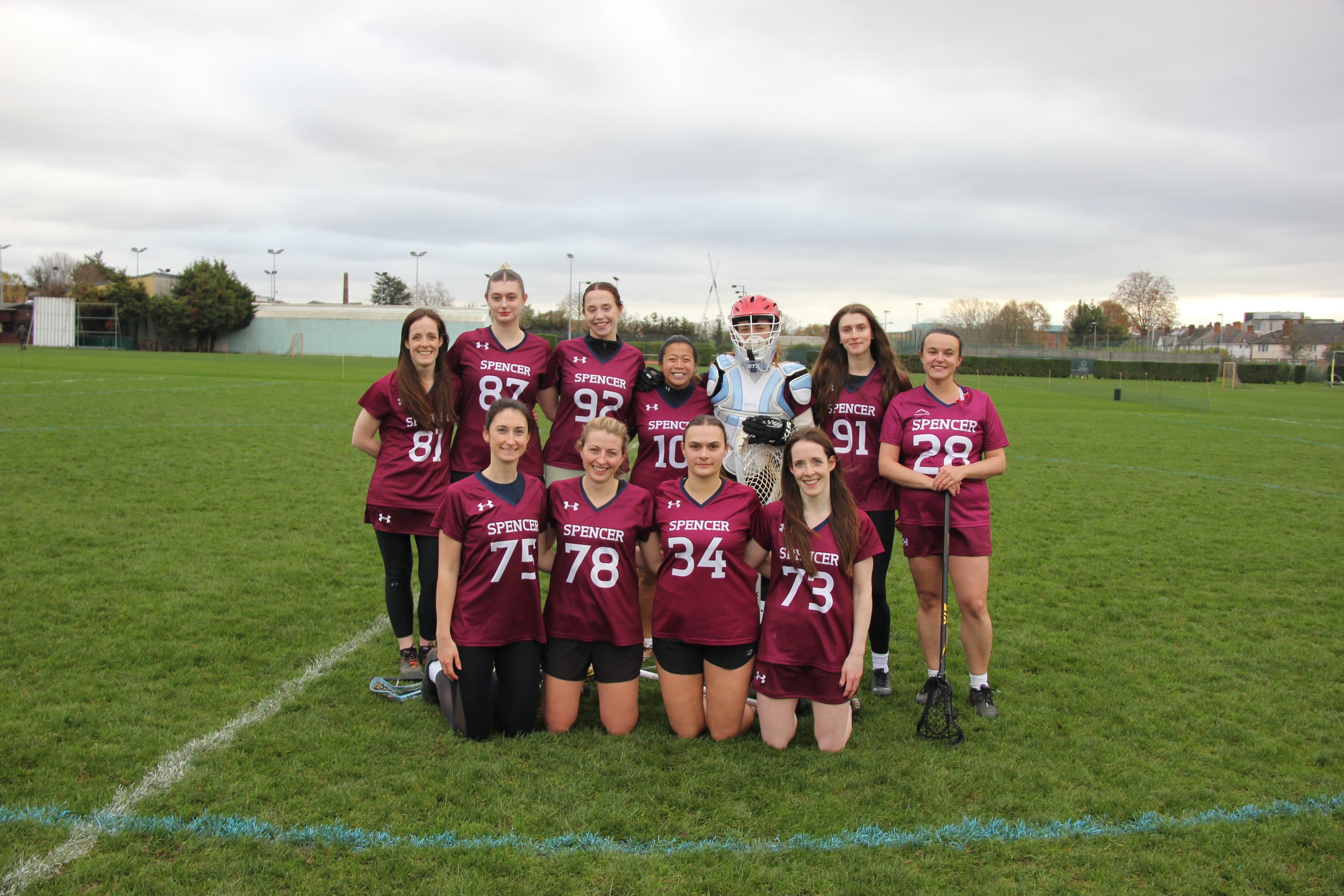 Lacrosse team of ten women in matching maroon jerseys with white numbers and 'SPENCER' on the front, posing on a grassy field with a goalie in protective gear, some women standing and some kneeling in front.