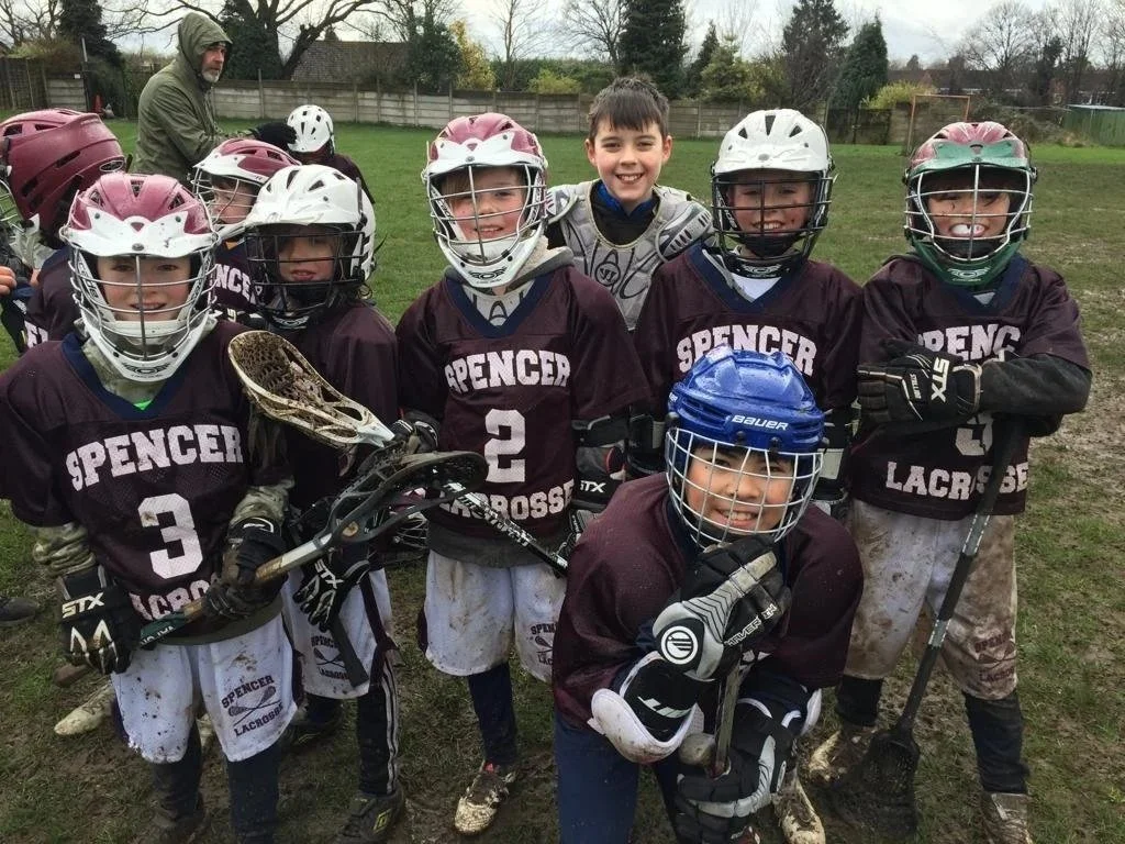 Young boys on a lacrosse team wearing maroon jerseys, helmets, and gloves, posing after a game on a muddy field with a coach or adult in the background.