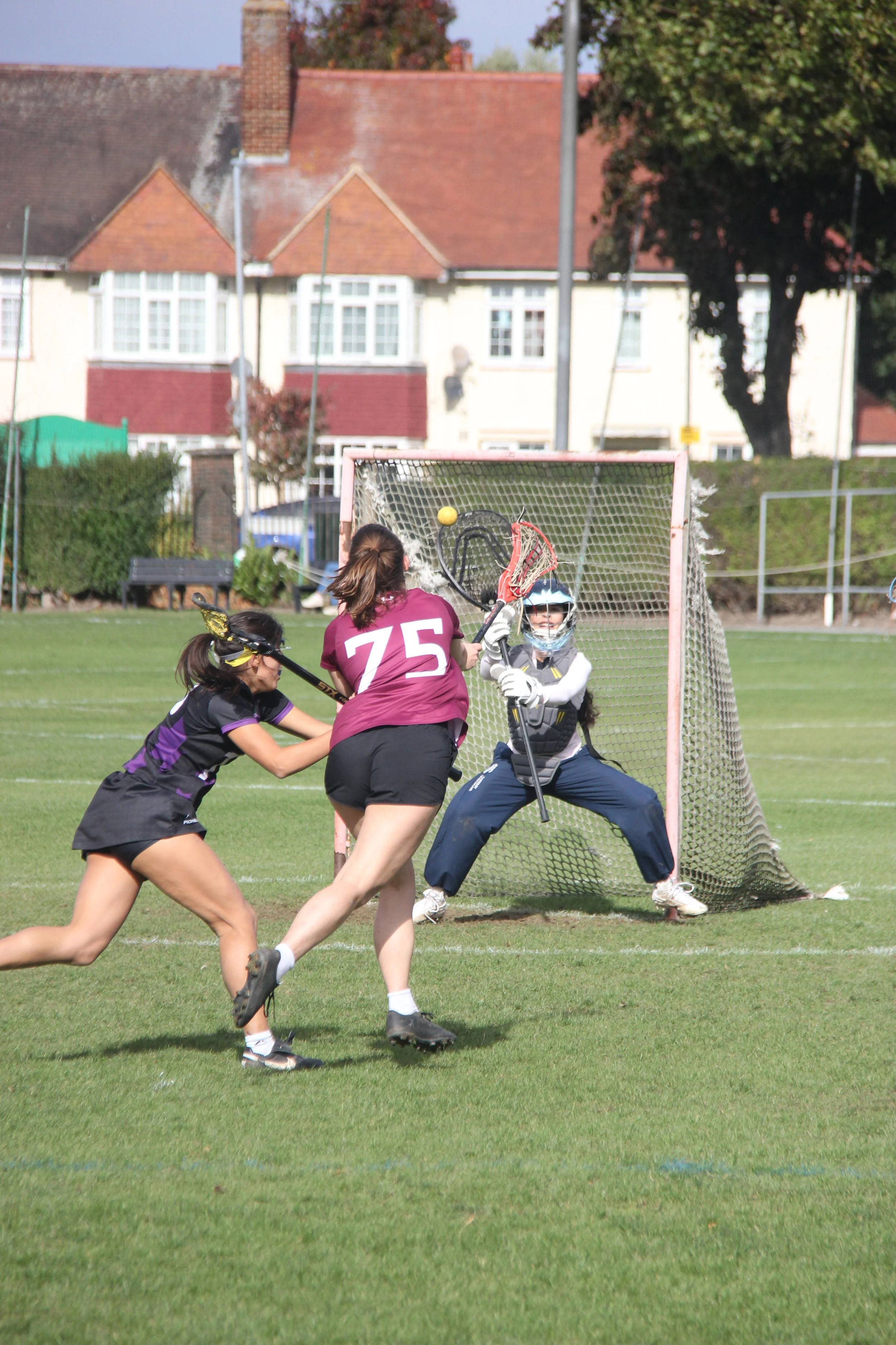 Two female lacrosse players contest a shot at goal as the goalkeeper attempts to block the shot on a grassy field in a residential neighborhood.
