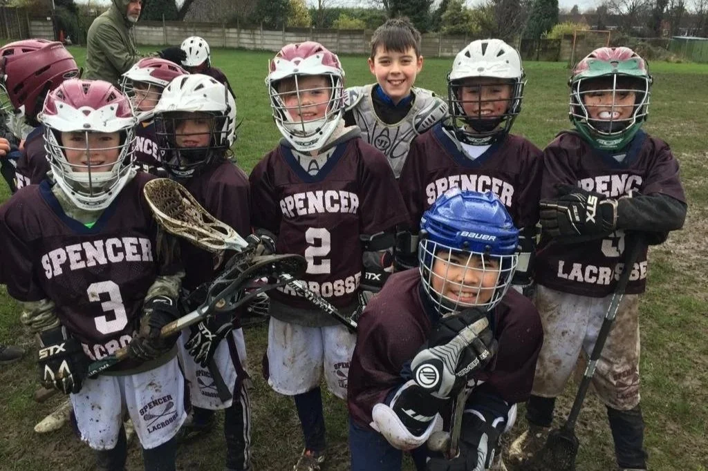 Young boys in lacrosse uniforms and helmets, outdoors on a grassy field, smiling for a team photo.