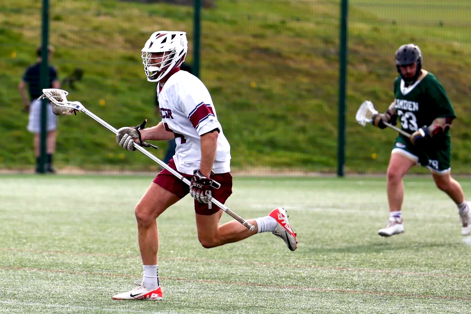 Lacrosse players in action on a field, one in a white and maroon uniform running with a lacrosse stick and wearing a helmet, while another in a green uniform runs behind him holding a lacrosse stick with a helmet, with a grassy hill and fence in the background.