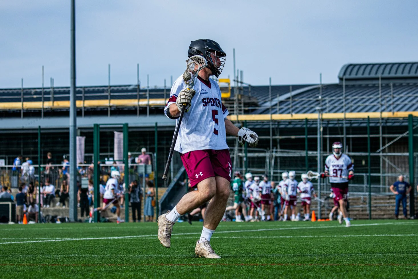 Lacrosse player in a white and maroon uniform running on a green field during a game with other players and spectators in the background.