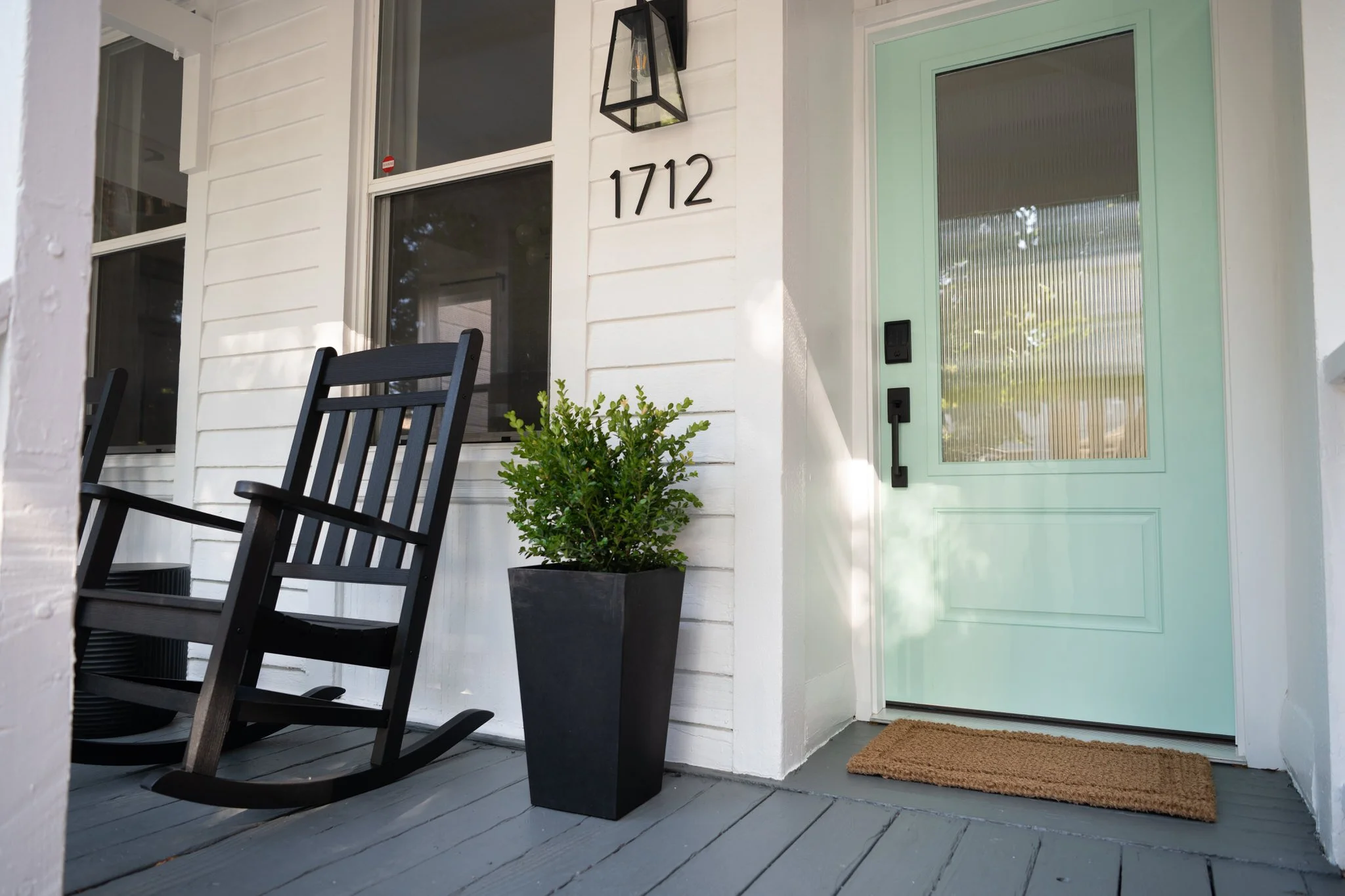 Front porch with a light blue door, black letterbox, and house number 1712, two black rocking chairs, a potted green plant, and a brown doormat.