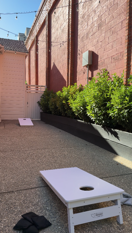 Outdoor cornhole game setup on a paved area next to a brick wall with green bushes. Cornhole boards are positioned for play, with one in the foreground and another in the background. A pair of black gloves rests on the ground near the closest board.