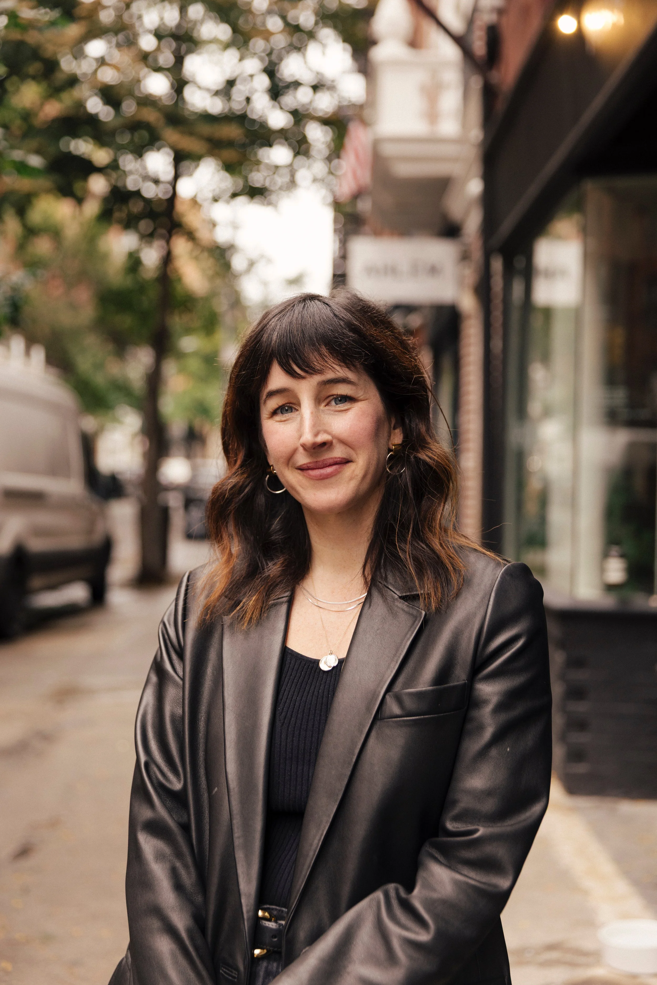 A woman with shoulder-length dark brown hair, wearing a black leather blazer over a black top, standing on a city sidewalk with trees and storefronts in the background.