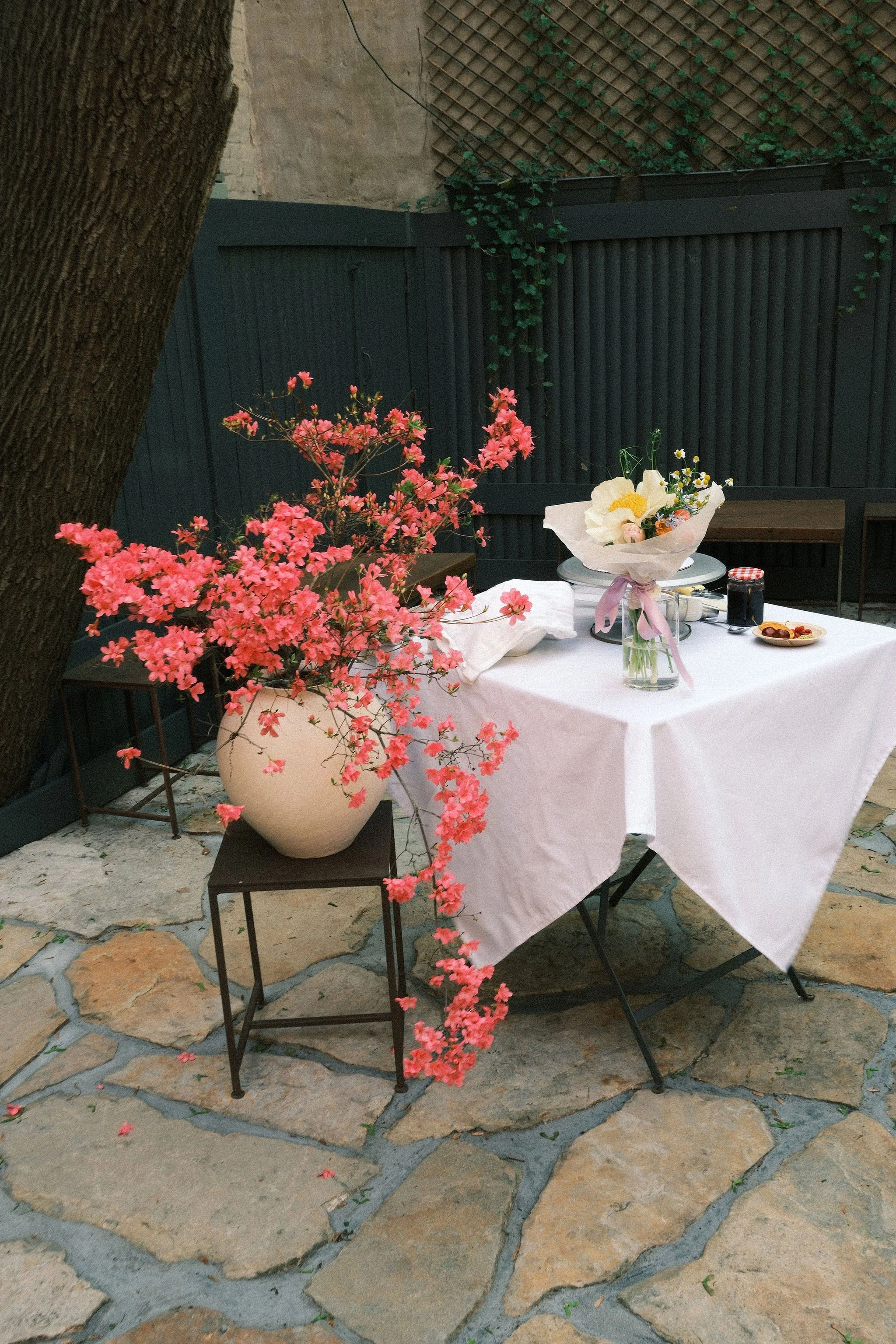 Outdoor dining table with a white tablecloth, decorated with a bouquet of flowers and some food, surrounded by pink flowers and a tree, set on a stone patio at dusk.