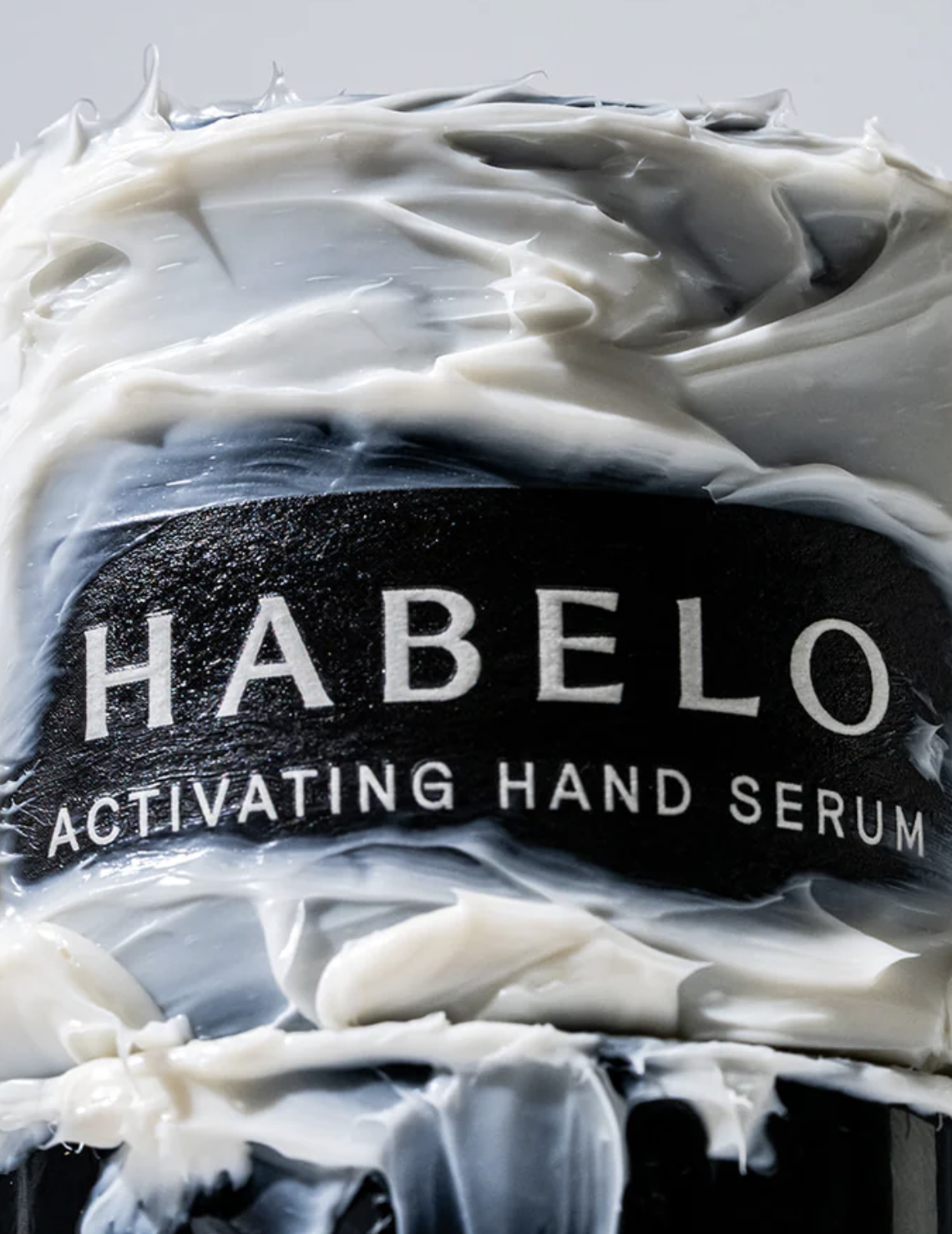 Close-up of a black container with the label 'HABELO ACTIVATING HAND SERUM' surrounded by smeared white cream or lotion.