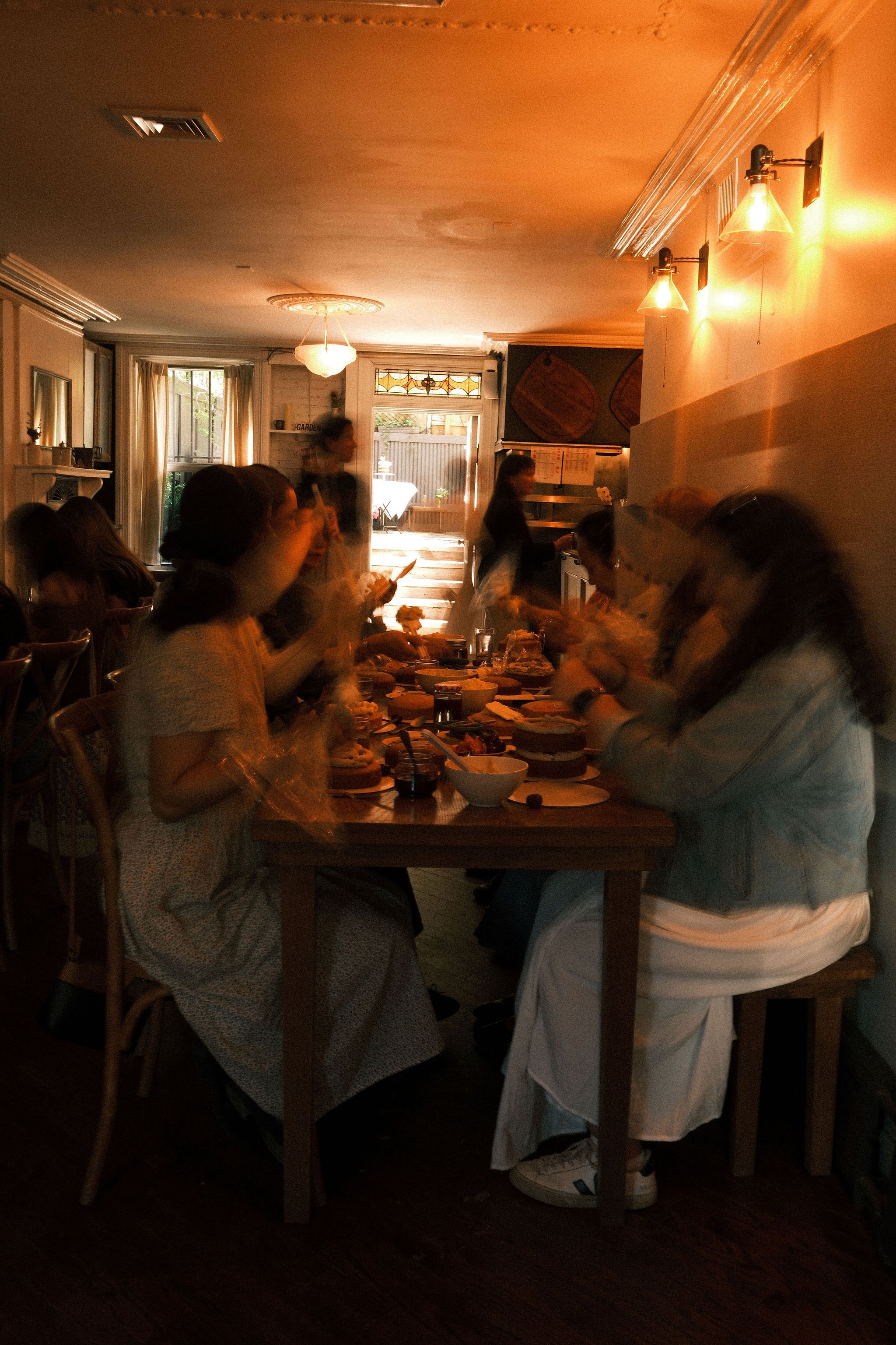 A group of people seated around a dining table in a cozy restaurant during evening time, with some waitstaff serving food, warm lighting, and a door leading to the outdoor patio.