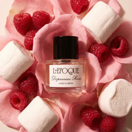 A bottle of L'EPOQUE Dopamine Rose perfume surrounded by raspberries, pink rose petals, marshmallows, and pink fabric.