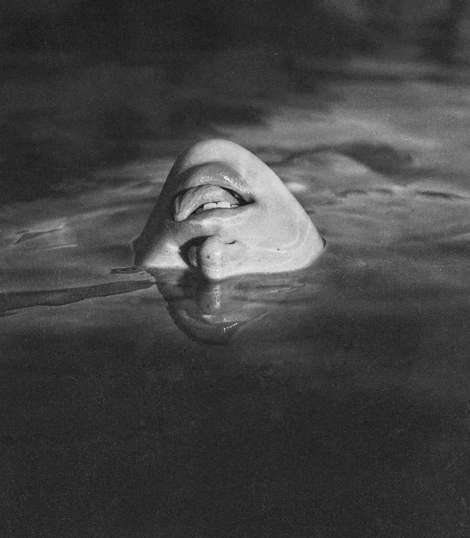Black and white photo of a person's face partially submerged in water, with only their mouth and part of their nose visible above the surface, showing a joyful expression.