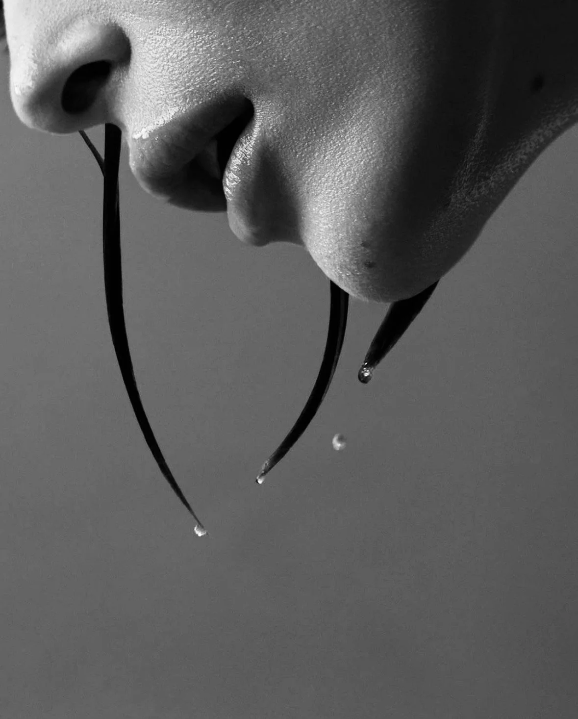 Close-up of a person's face in profile with water droplets on their lips and chin, water dripping from two black strings held near their lips, black and white photo.