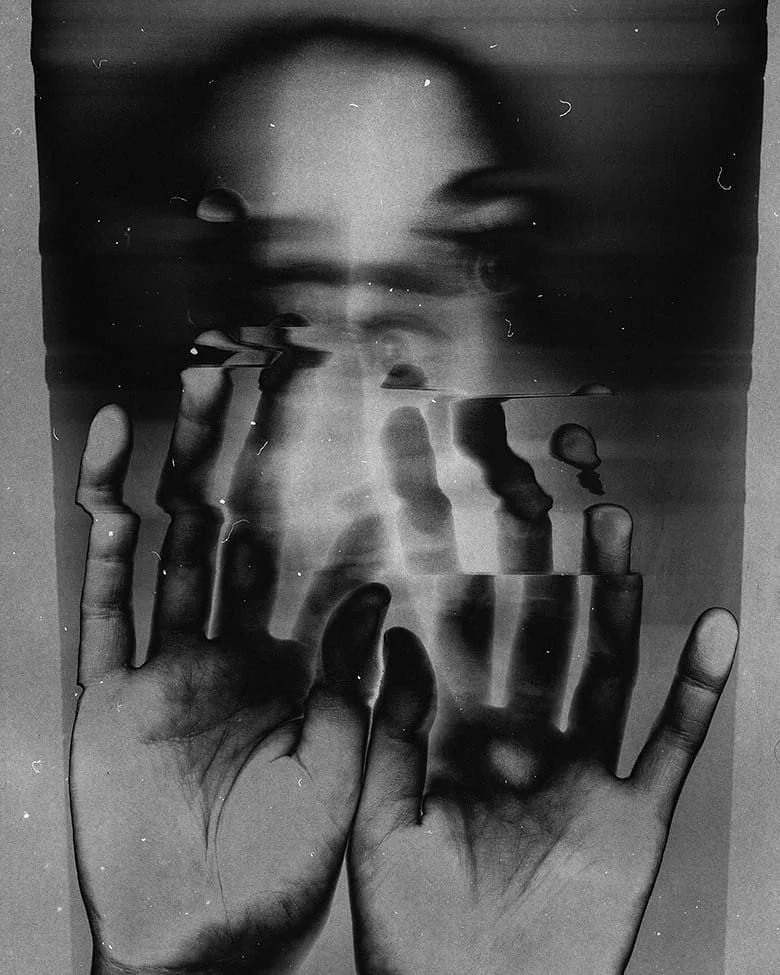 A person's face viewed through a glass surface, with hands pressed against the glass, creating a distorted, reflective effect in black and white.